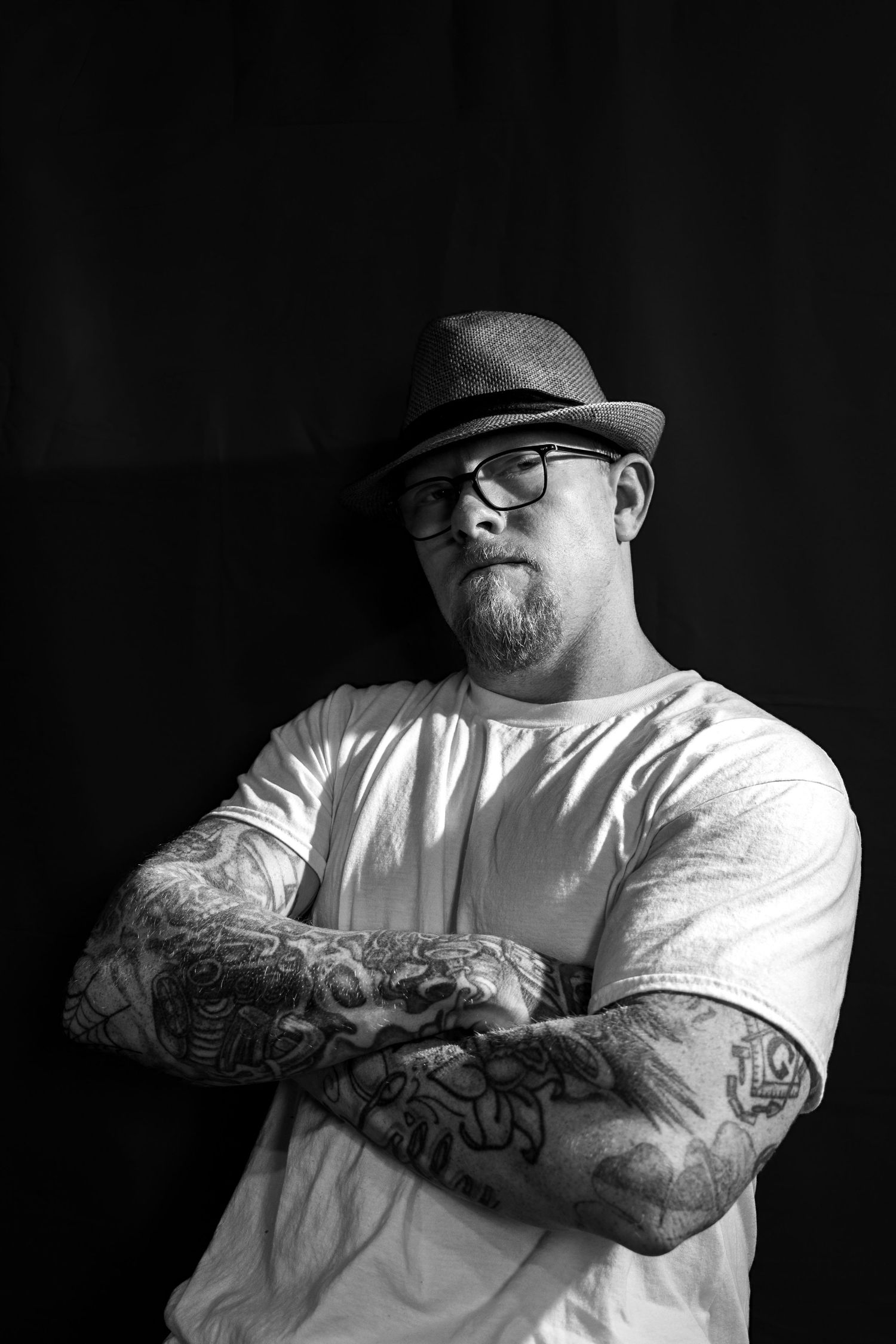 Black and white portrait of tattooed man in fedora and glasses with arms crossed against dark wall.