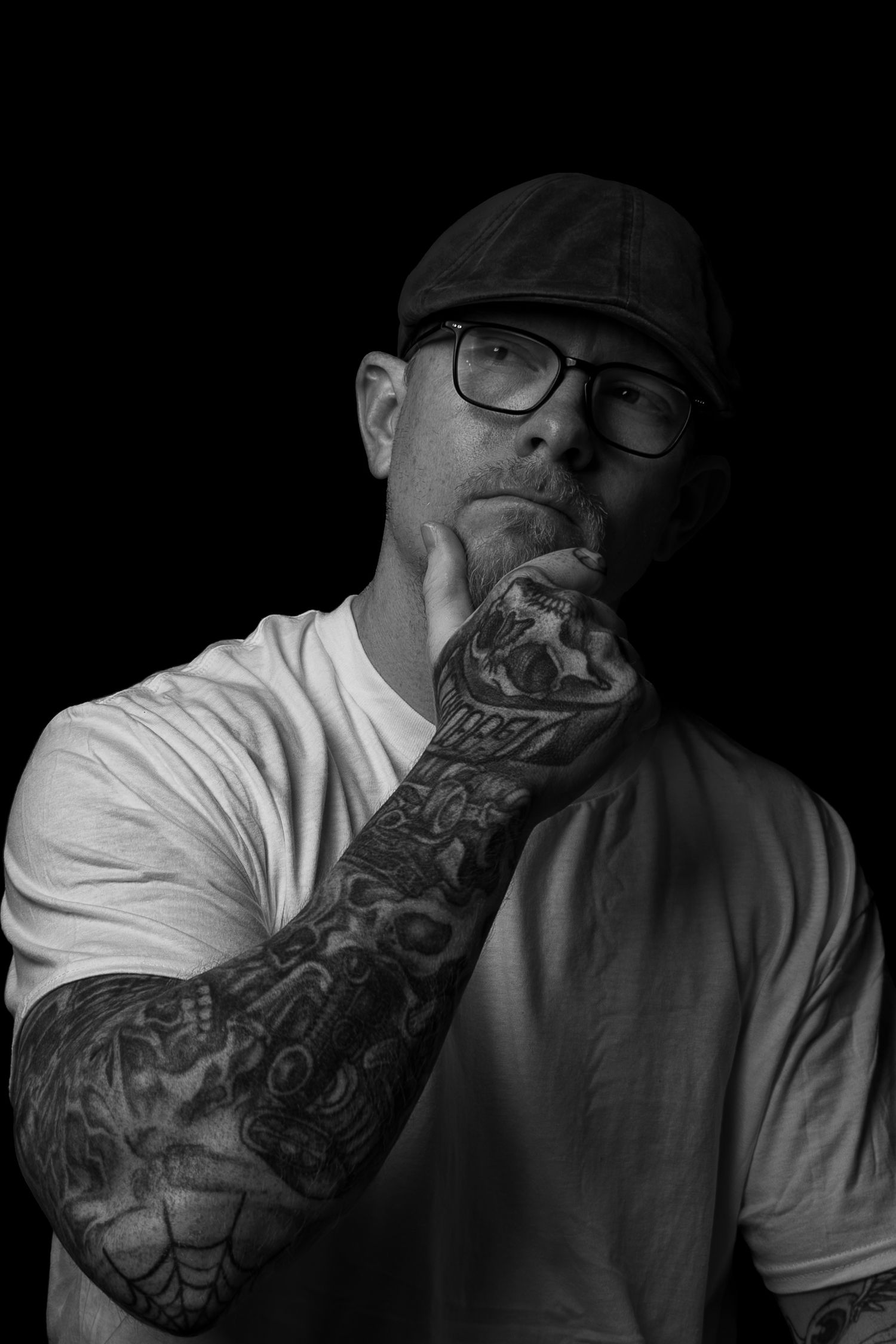 Black and white portrait of a tattooed man in a flat cap and glasses thoughtfully resting his chin on his inked hand.