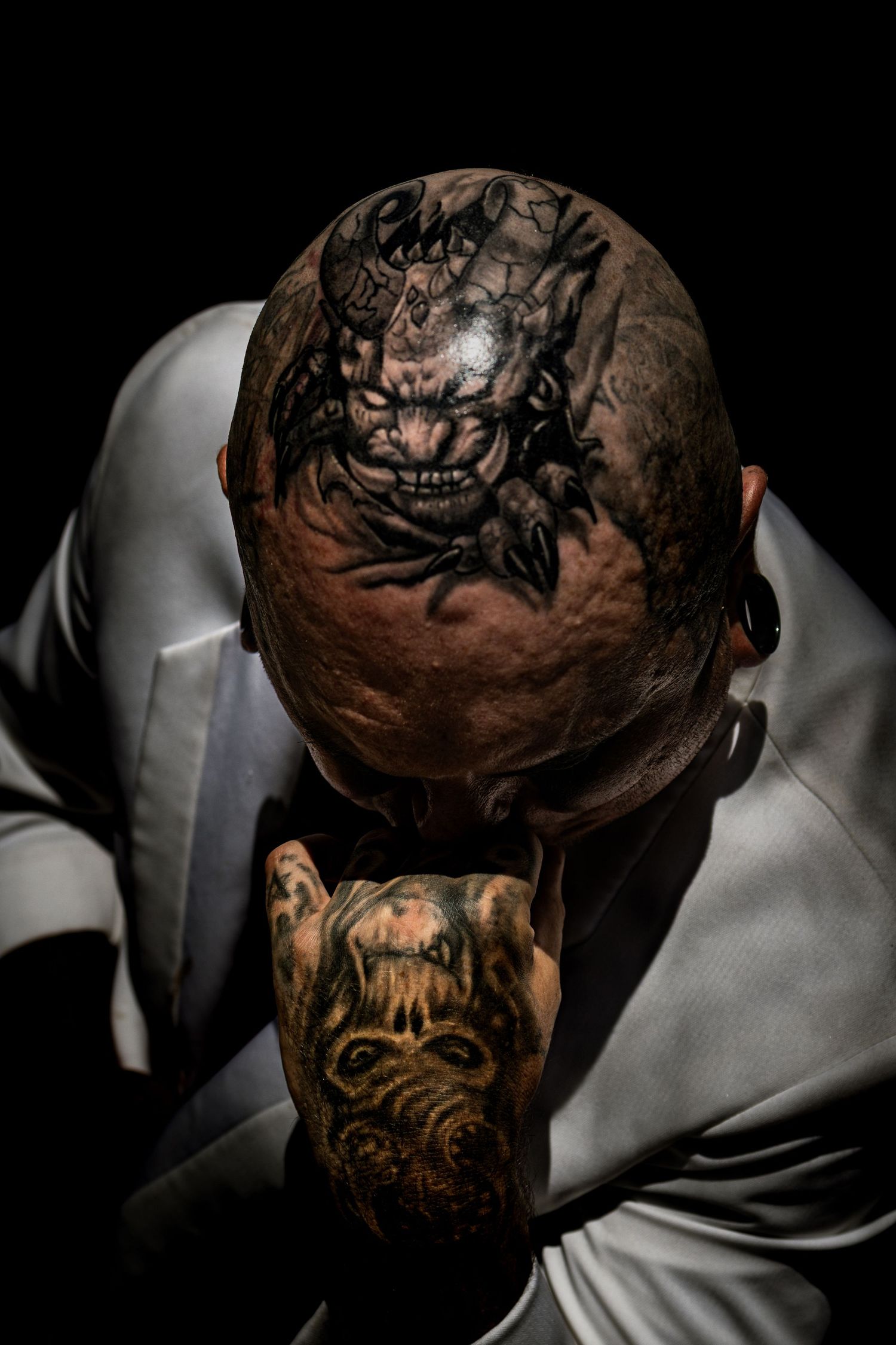 Close-up portrait of a bald man with intricate horror-themed tattoos covering his head and hand against dark background.