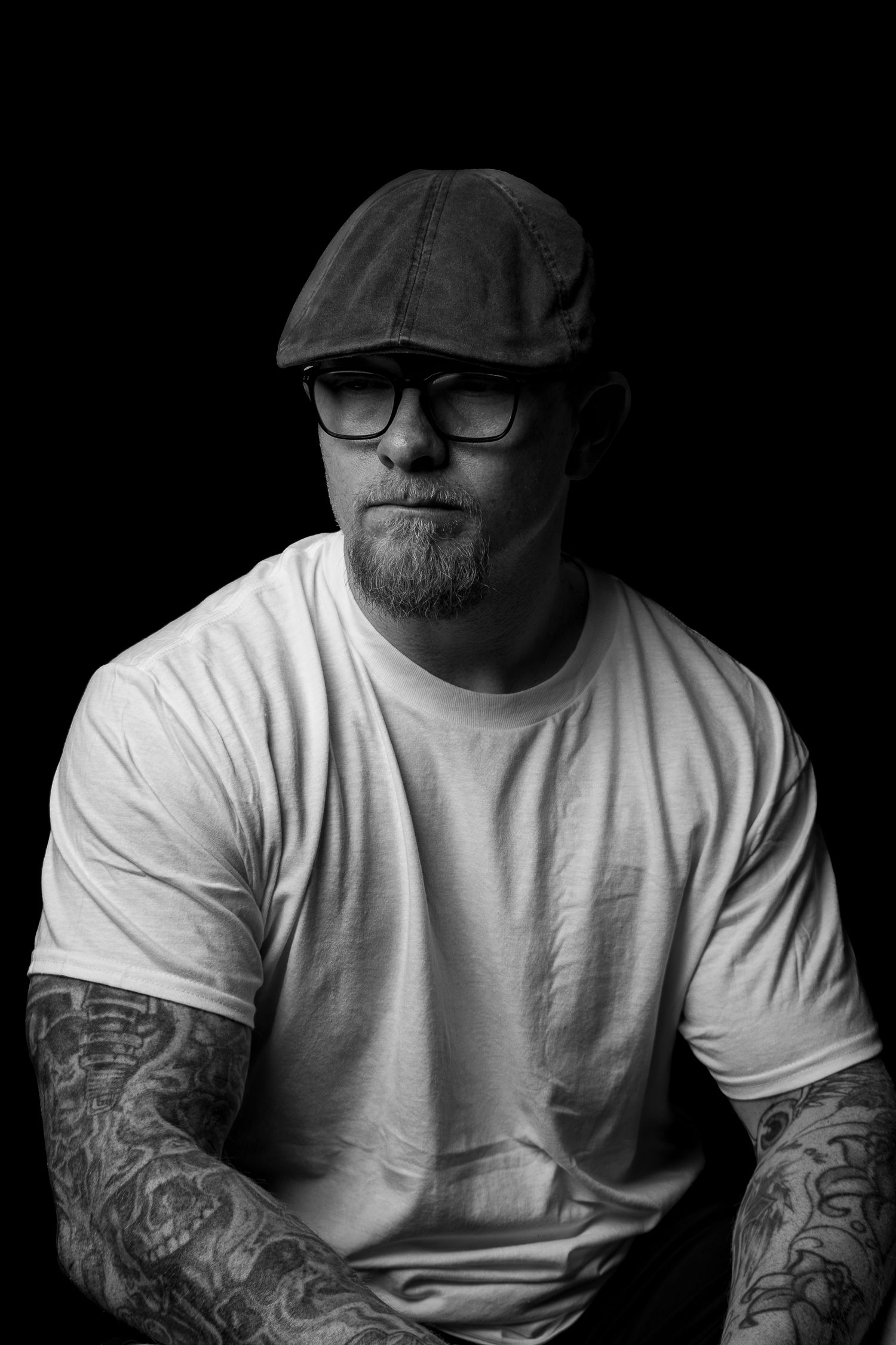 Black and white low-angle portrait of tattooed bearded man in cap and glasses on dark background.