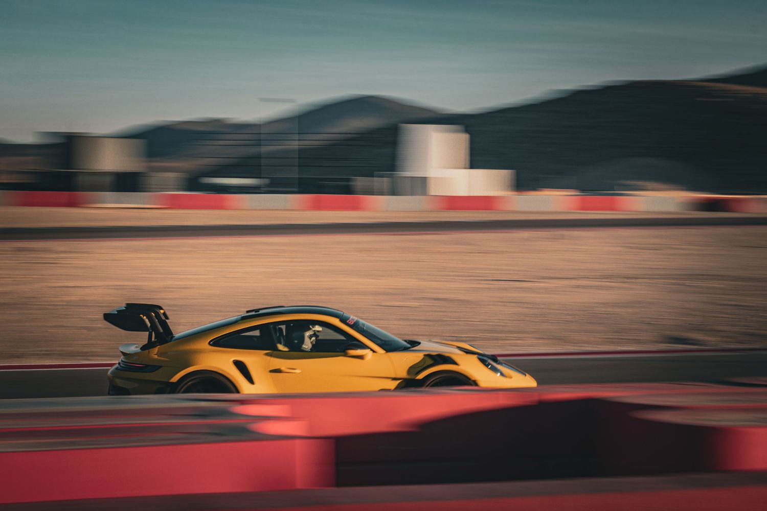 Yellow Porsche 911 GT3 RS speeding on a race track with motion blur effect and mountains behind.