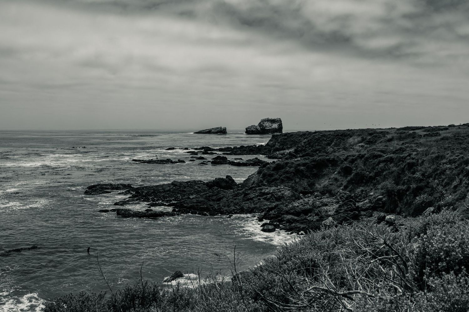 Black and white rocky coastal cliff with crashing waves and a ruined structure under dramatic overcast skies.