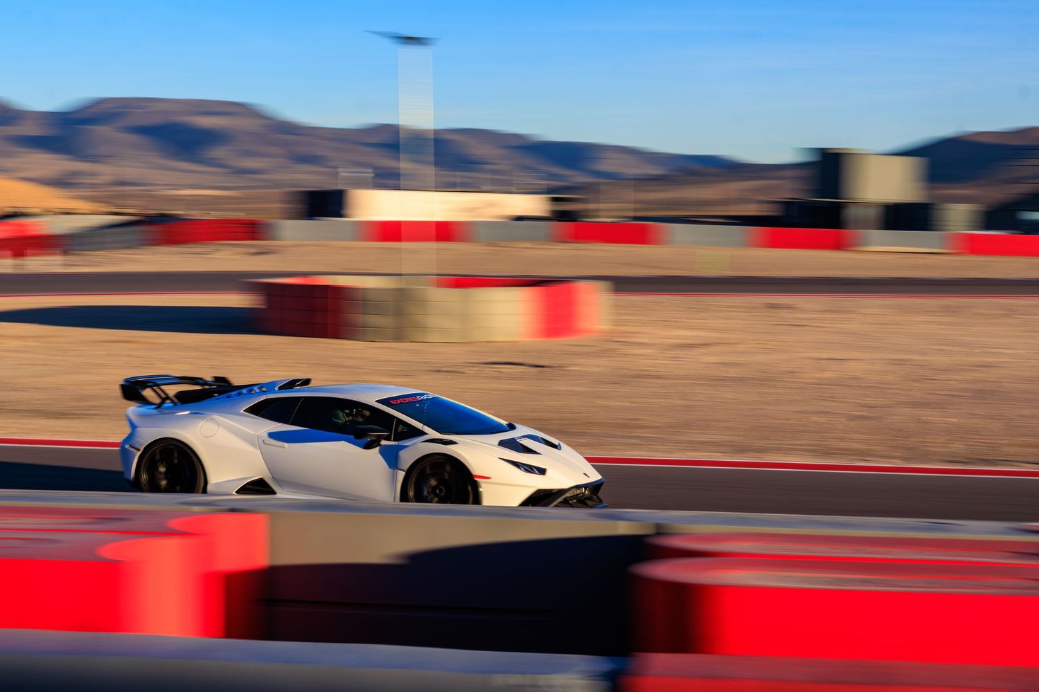 White high-performance track car with rear spoiler speeding past red barriers on a desert racing circuit.