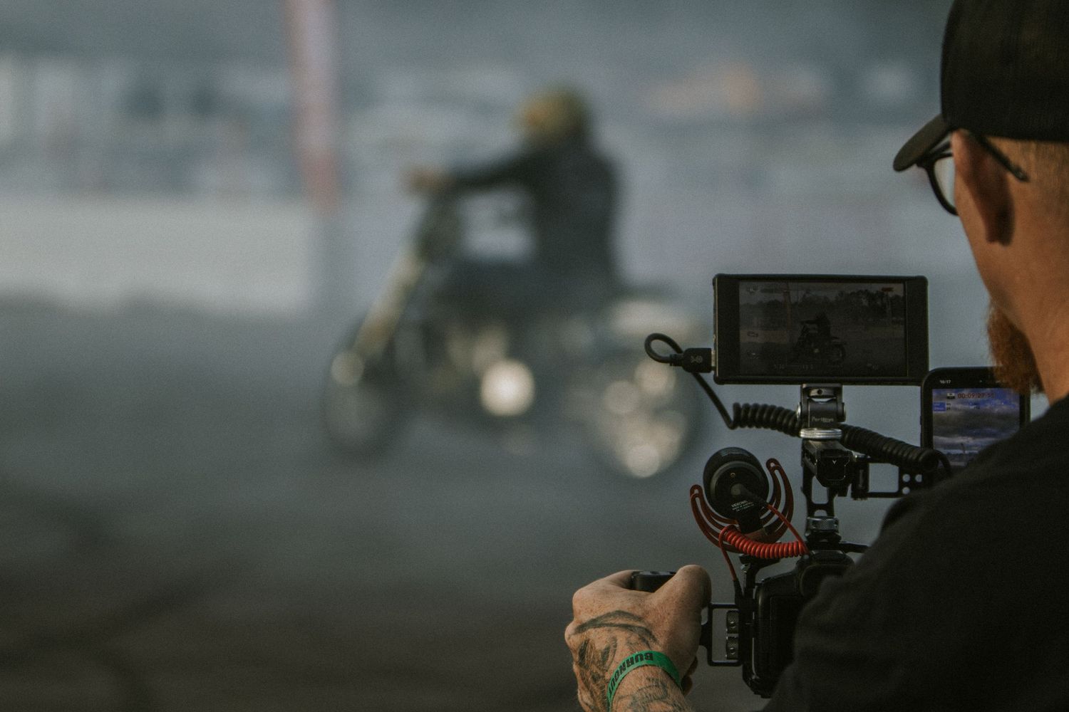 A tattooed filmmaker captures footage of a motorcyclist performing stunts in a smoky outdoor arena.