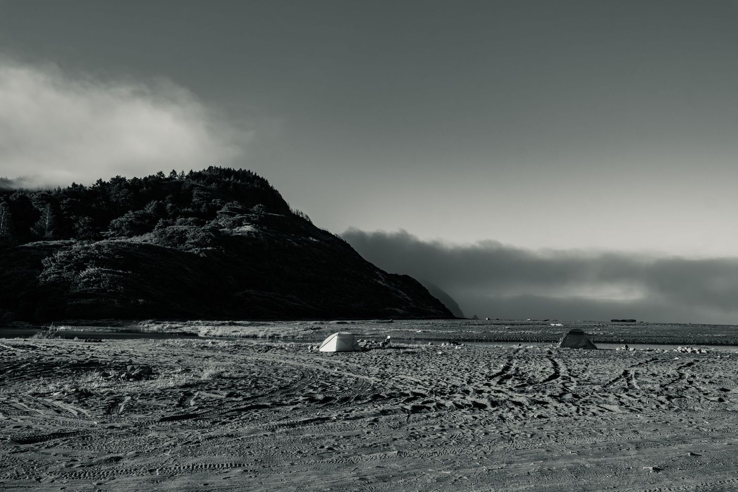 Black and white beach scene with two small tents on sandy shore beside a large misty mountain under cloudy skies.