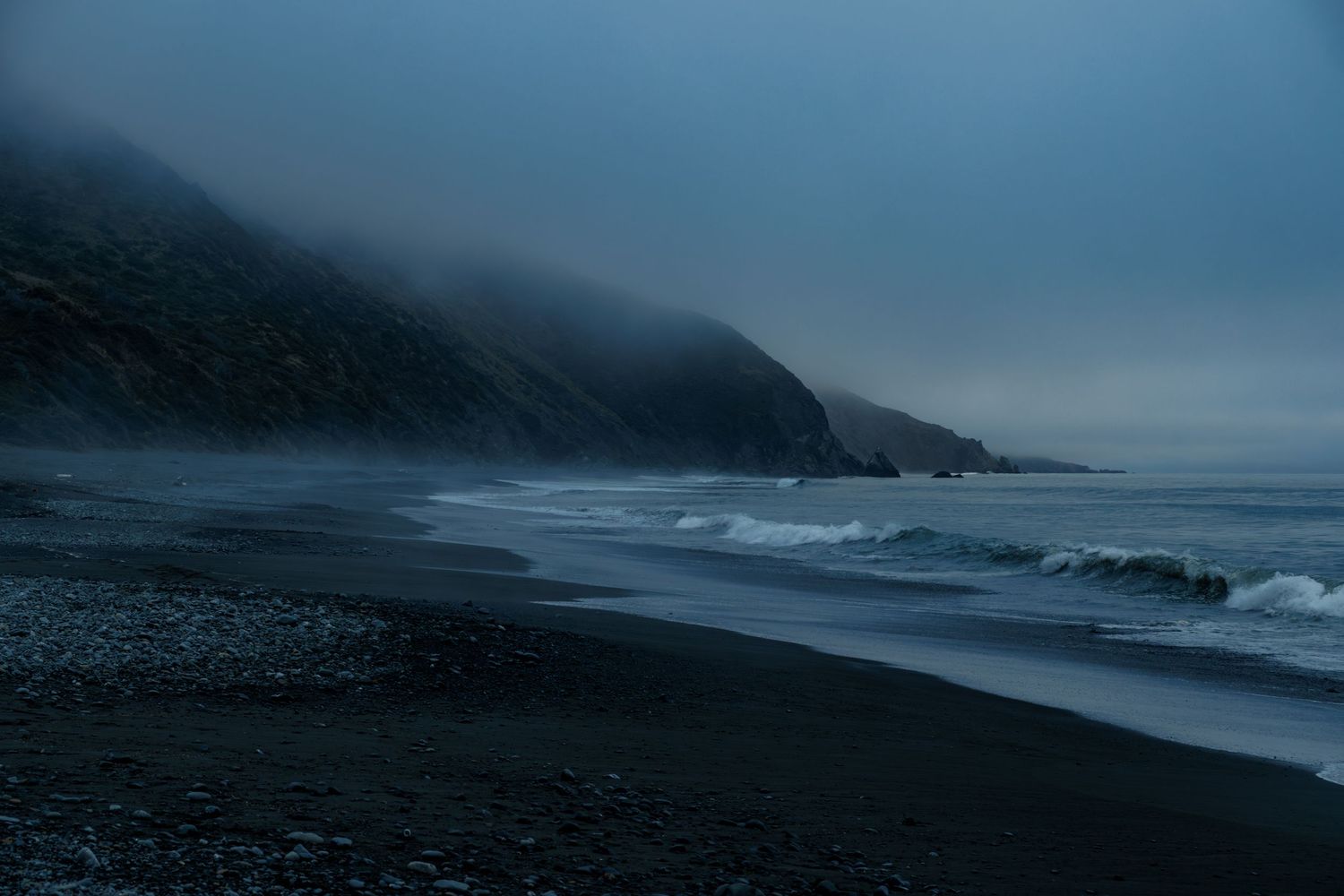 A moody dark blue seascape with misty mountains and gentle waves rolling onto a rocky beach shore.
