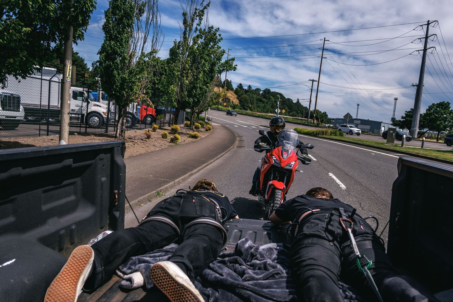 View from a truck bed of a red motorcycle riding on a tree-lined road under a partly cloudy sky.