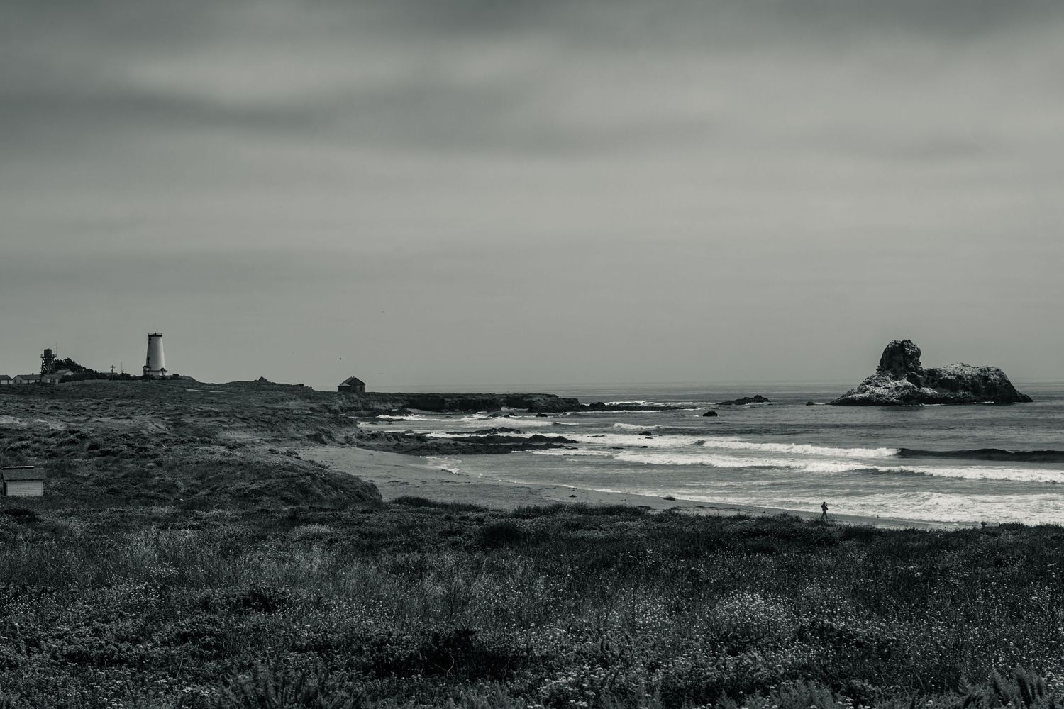 Black and white coastal landscape with rocky shoreline, crashing waves, and a lighthouse under overcast skies.