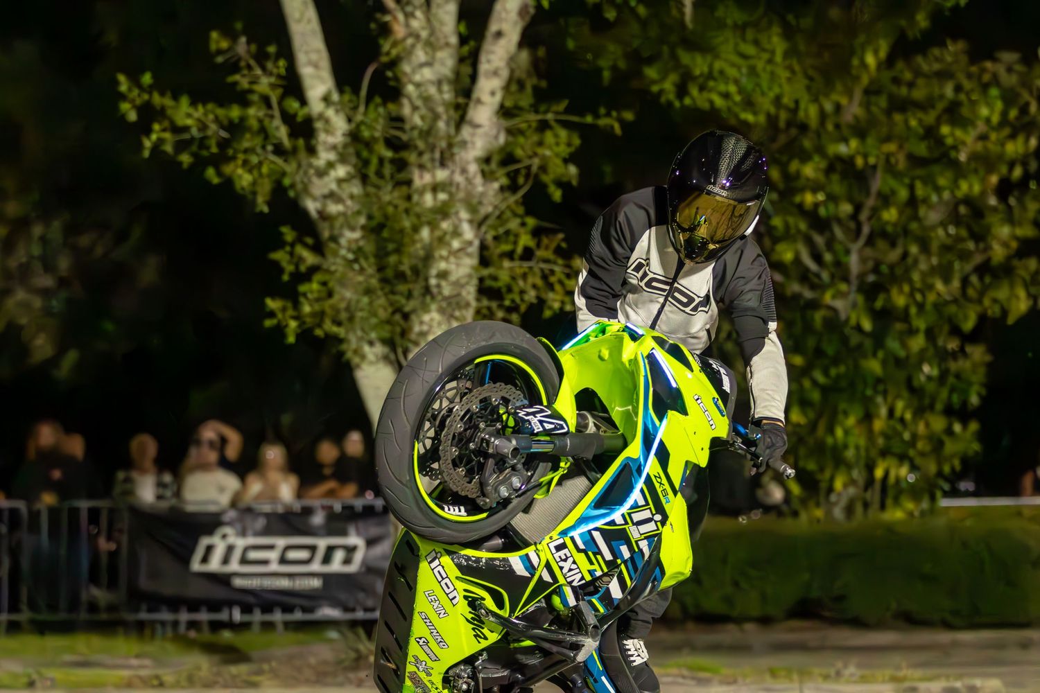 Motorcycle stunt rider performing a wheelie on a neon green bike at a nighttime show.