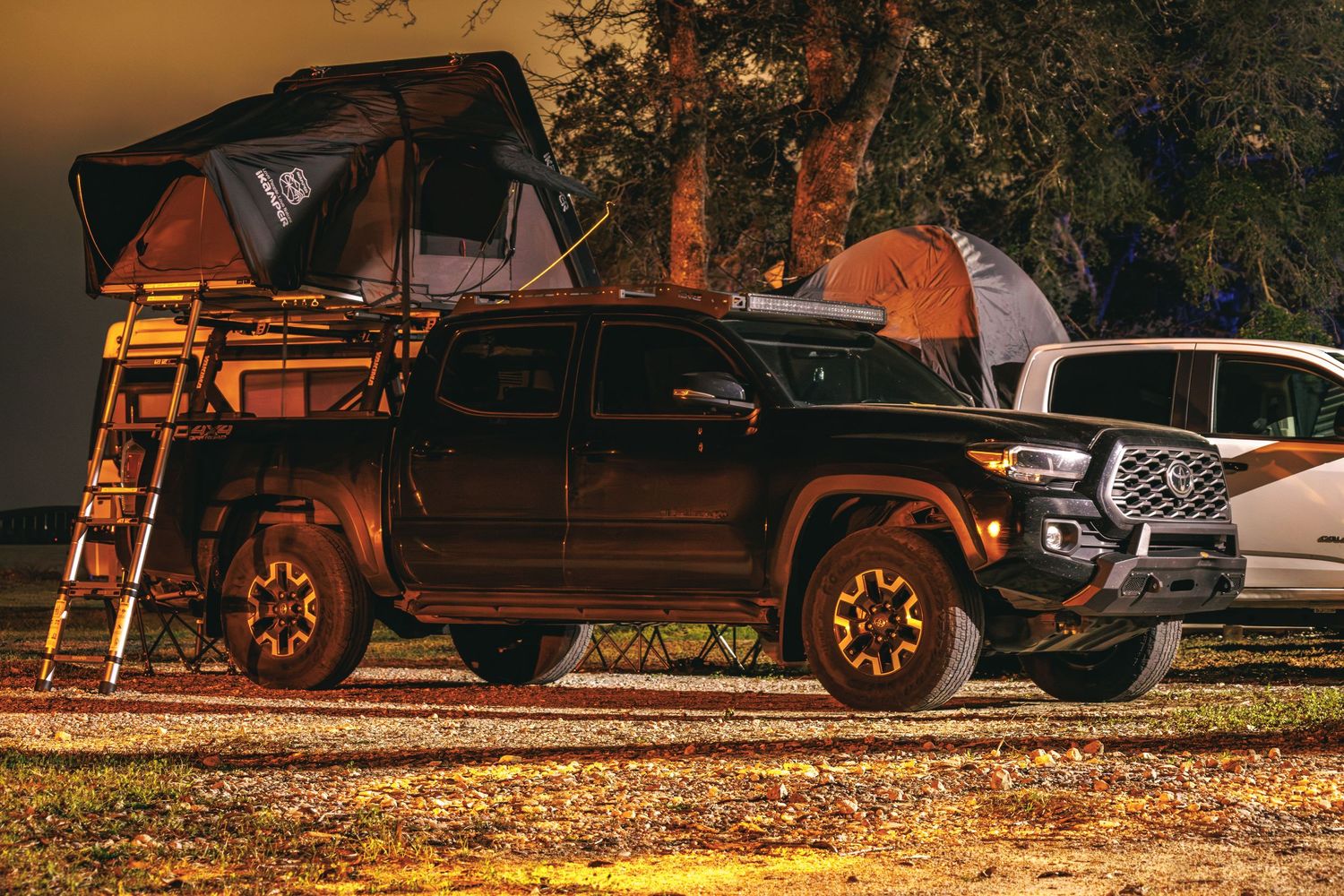 Black Toyota Tacoma with rooftop tent at a campground illuminated by warm lights.