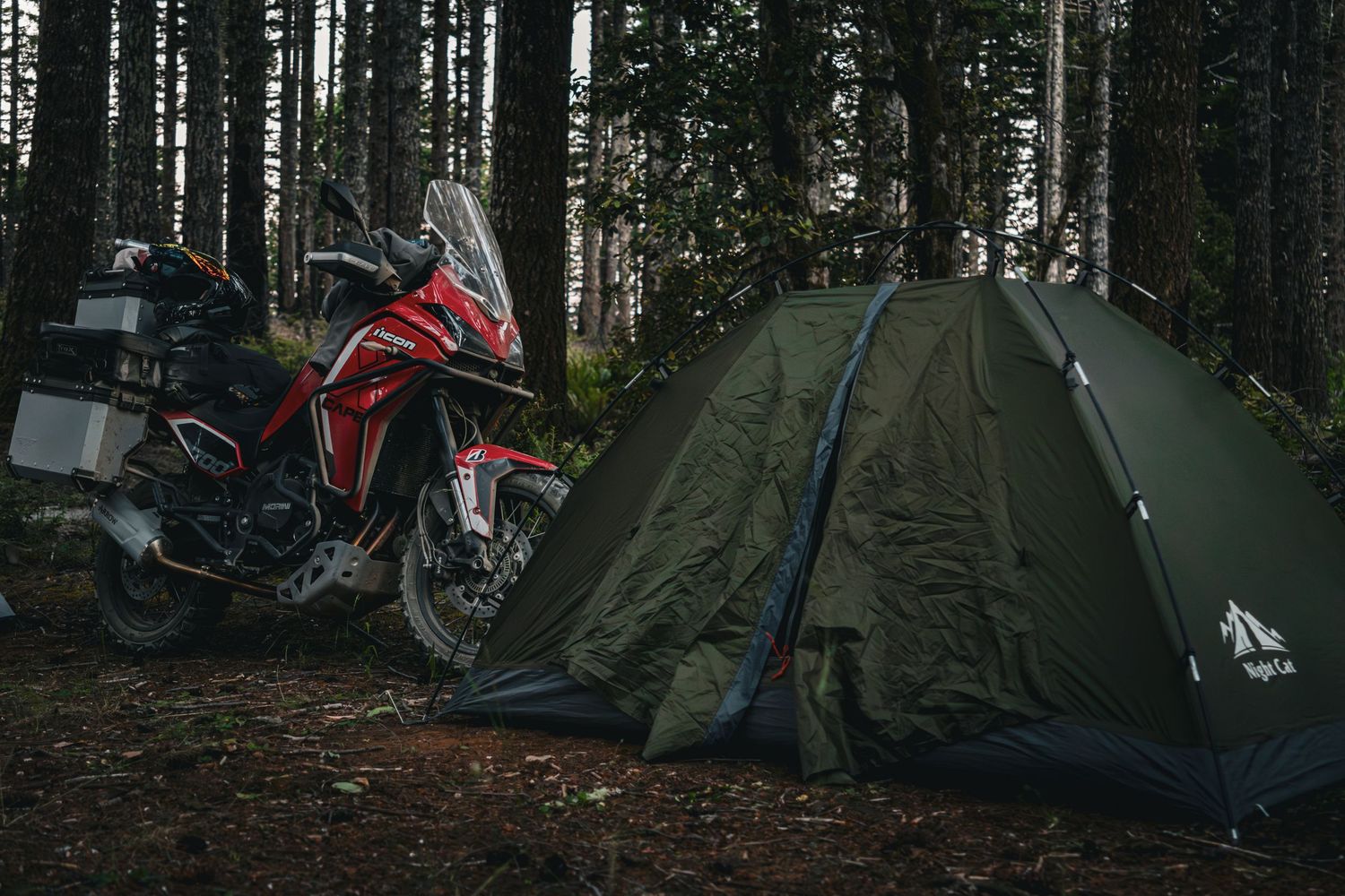 Red adventure motorcycle parked beside a green camping tent in a dense forest setting.