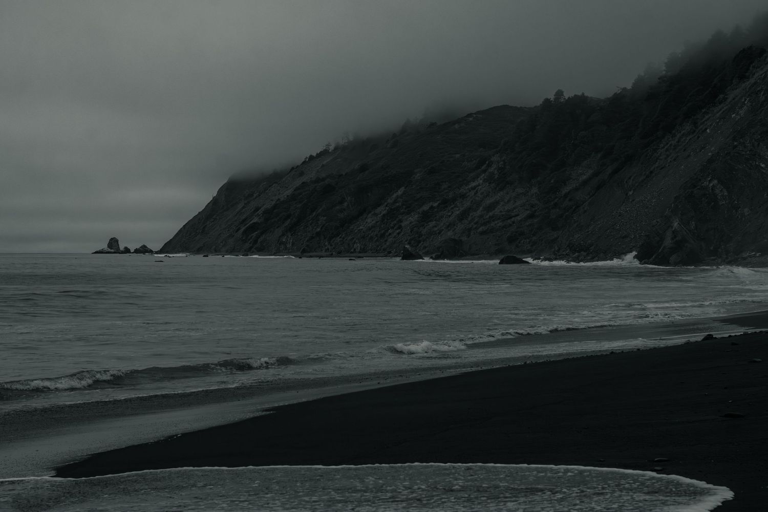 Dark moody black and white seascape featuring a steep misty cliff and black sand beach with gentle waves.
