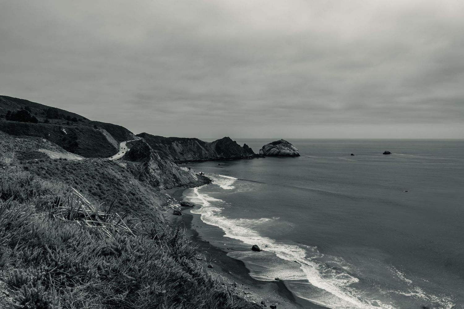 Black and white coastal landscape with winding road along dramatic cliffs and rocky shoreline under overcast skies.