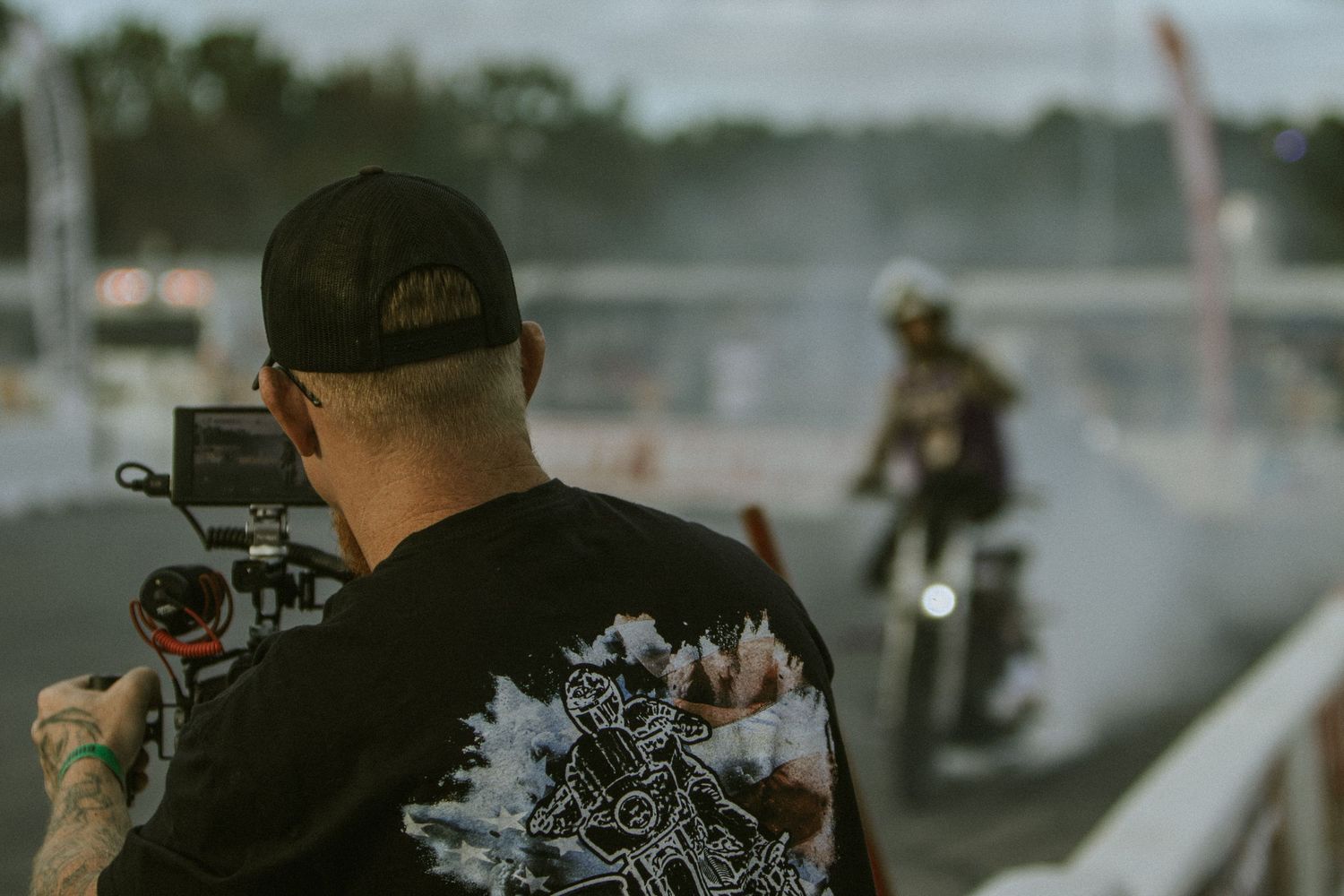 Tattooed cameraman filming a motorcycle stunt rider performing burnout on a track at dusk.