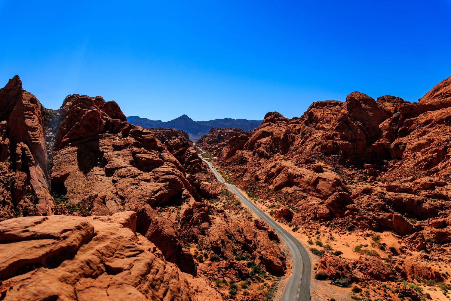 Aerial view of winding road cutting through dramatic red rock canyon formations under clear blue sky.