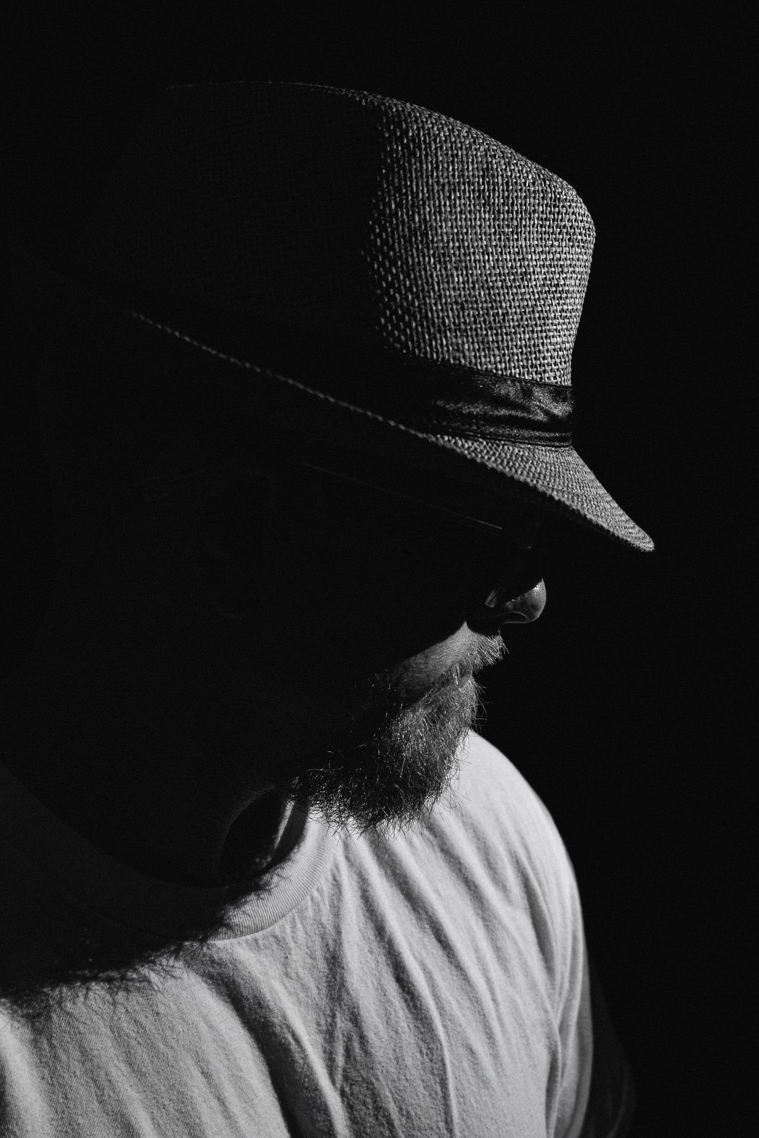 Dramatic black and white portrait of a bearded man wearing a fedora hat against dark background.