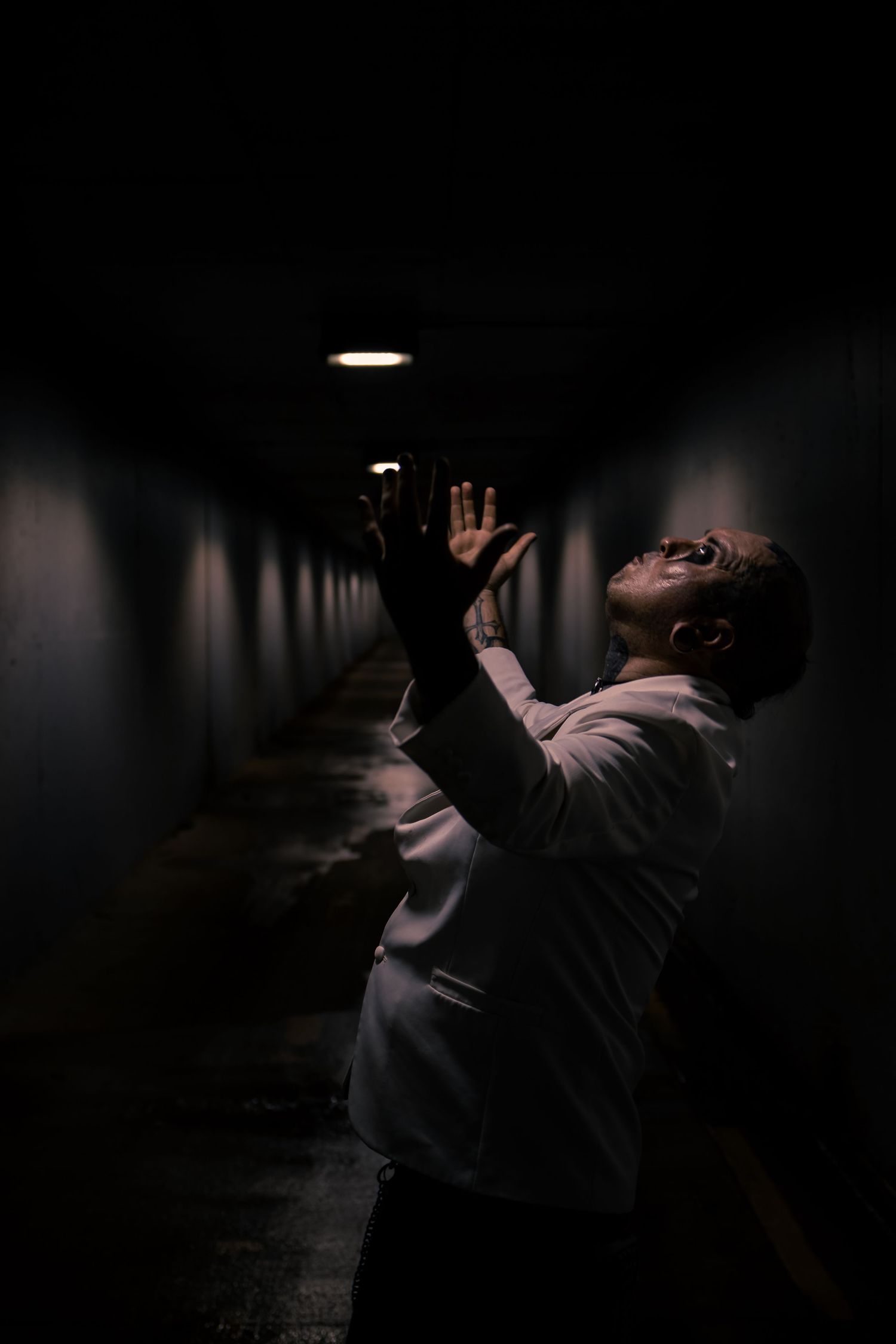 Dramatic low-light photo of a man reaching upward in a dark narrow corridor illuminated by a single overhead light.