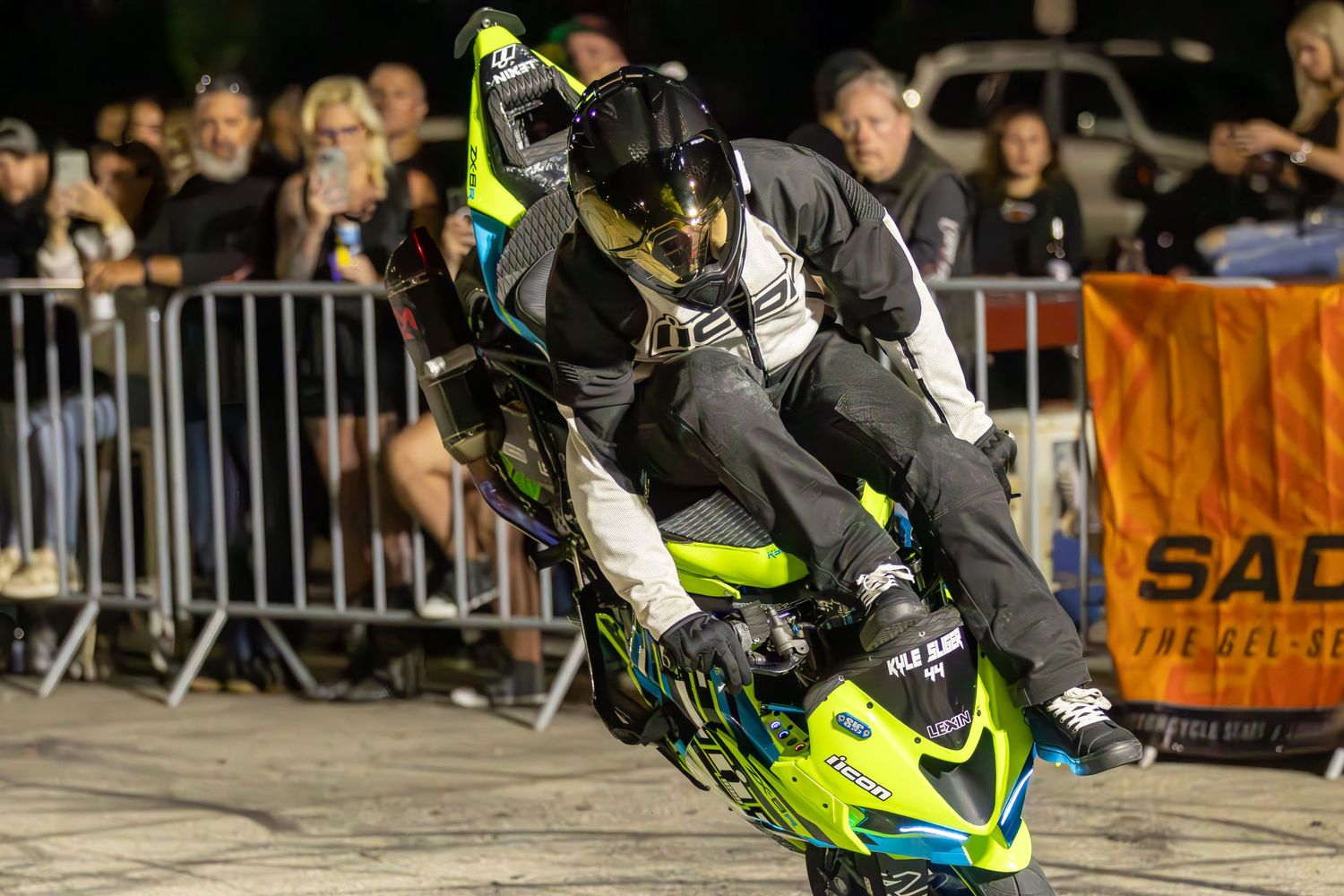 Stunt rider performing an extreme wheelie on a neon green motorcycle before a nighttime crowd.