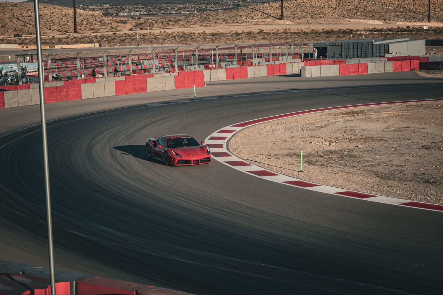 Red sports car navigating a curved asphalt racing track at a desert circuit with red barrier walls.