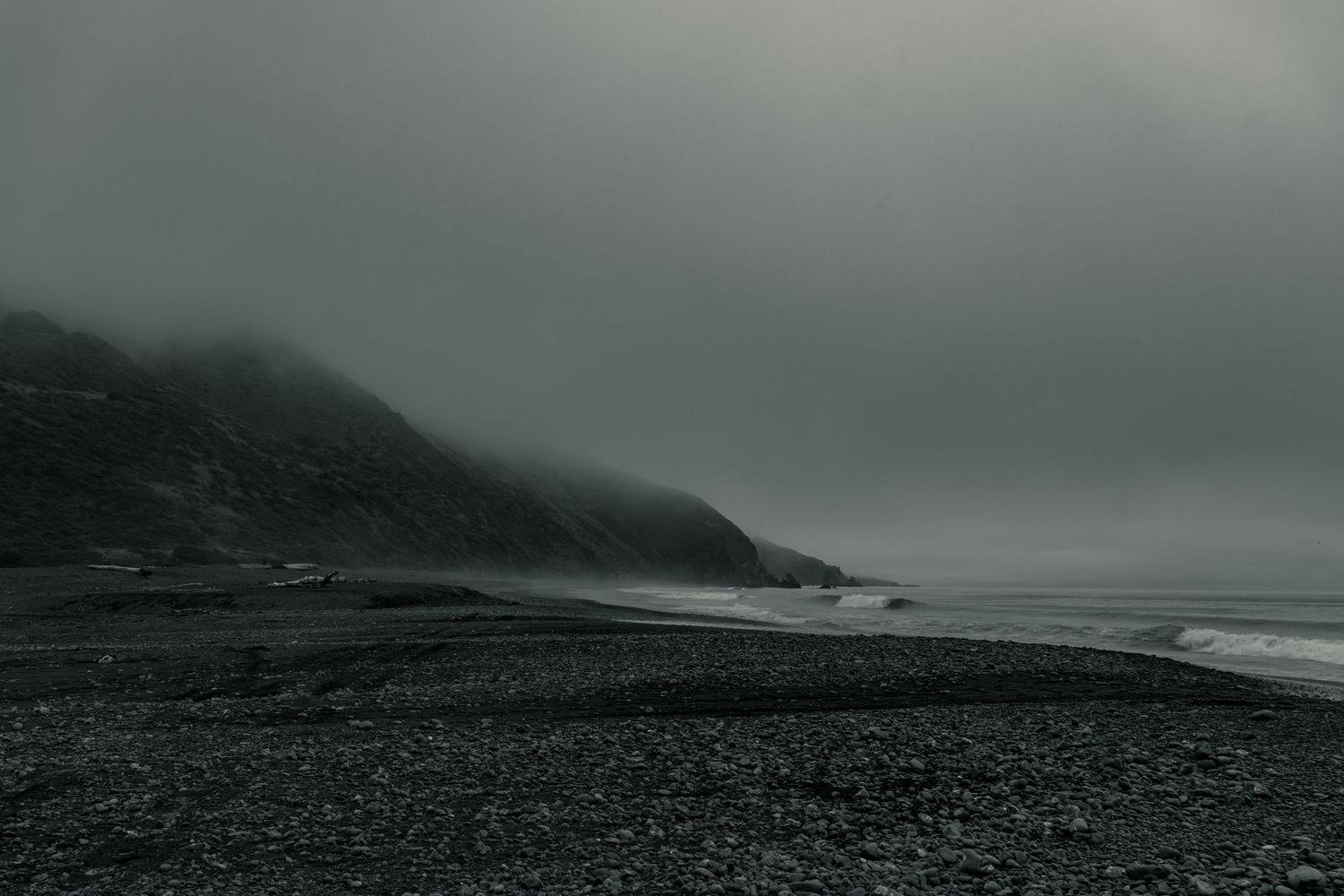 Moody black and white coastal landscape with rocky beach, misty cliffs, and overcast sky.