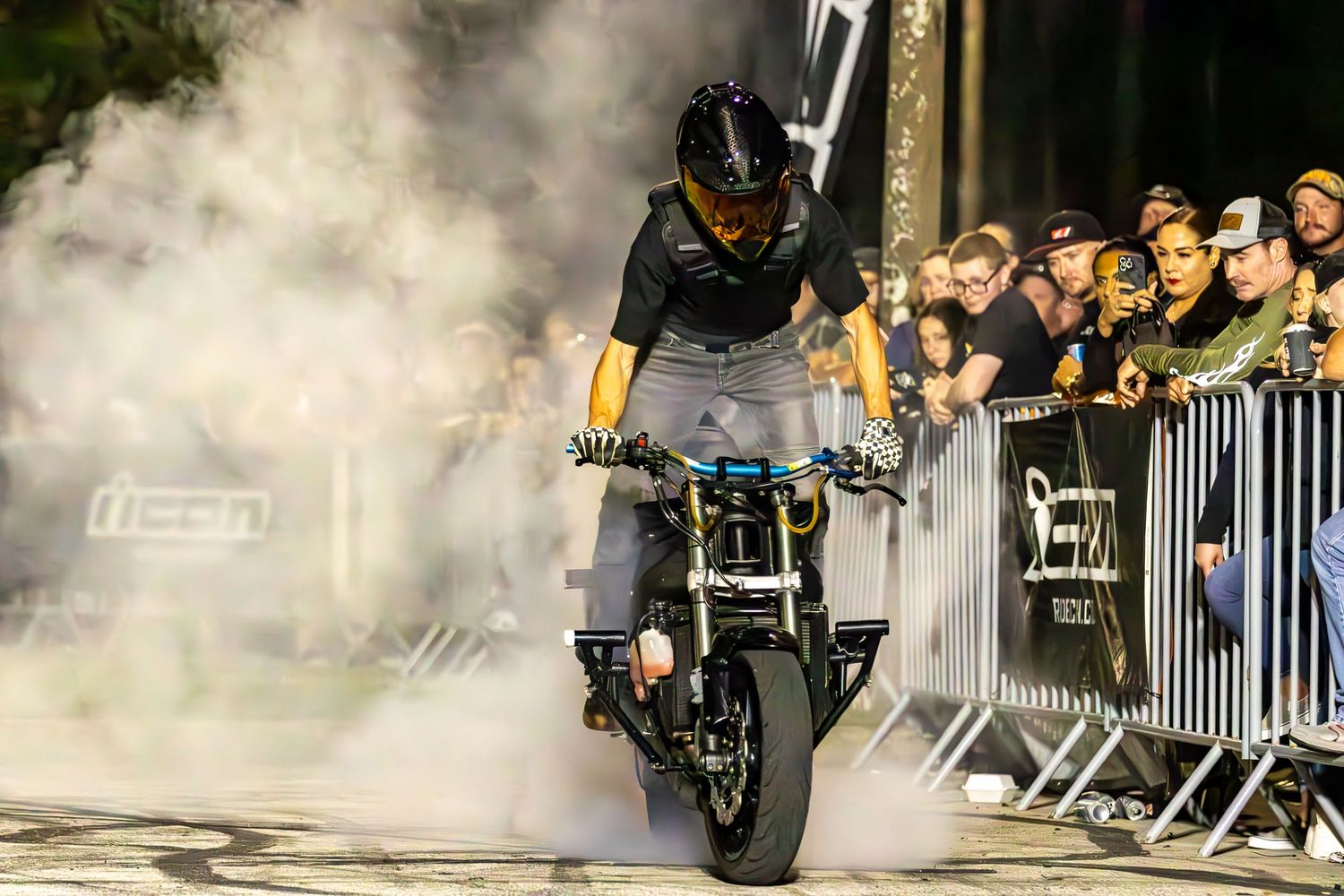Stunt motorcyclist performing a burnout in smoky arena with crowd watching behind barriers.
