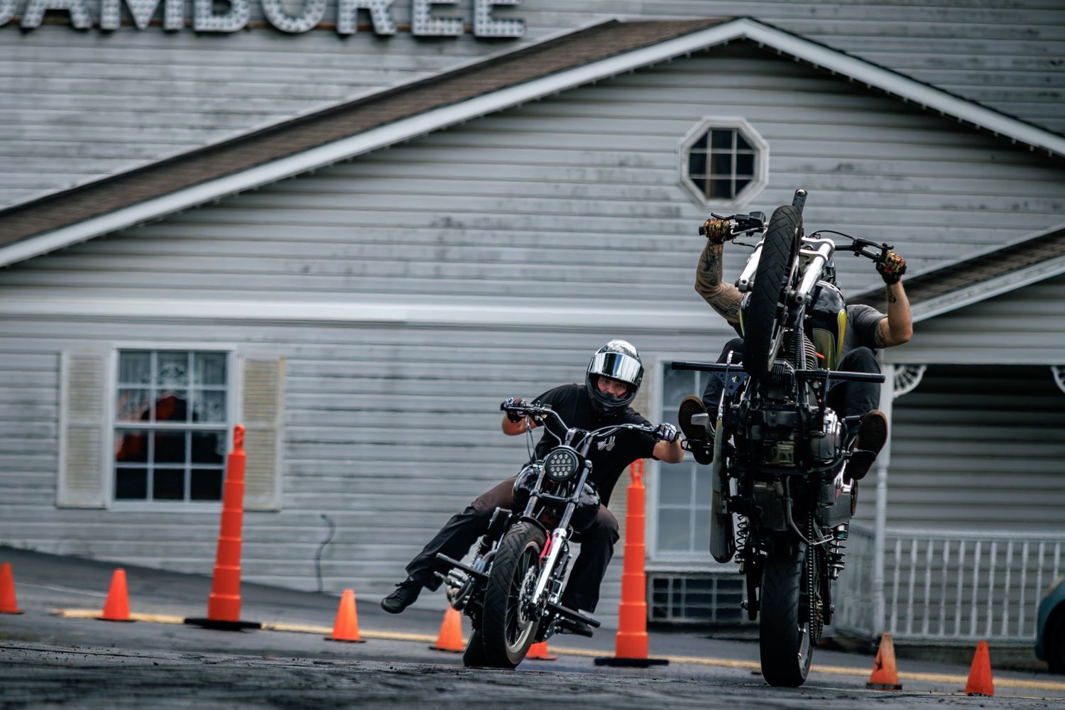 Two motorcyclists performing stunts near orange cones outside a Jamboree building on a wet road.