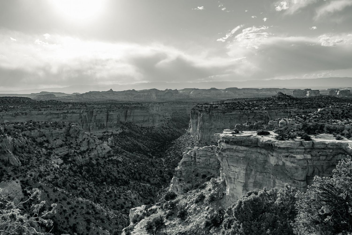 Black and white aerial view of a dramatic canyon landscape with mesas and cliffs under a sunlit cloudy sky.