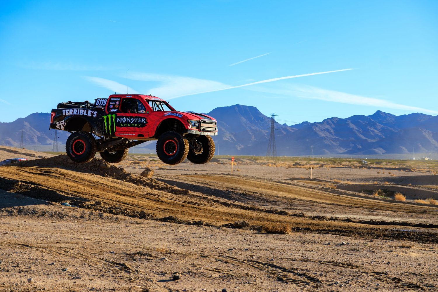 Red Monster Energy trophy truck catching massive air during an off-road desert race event.