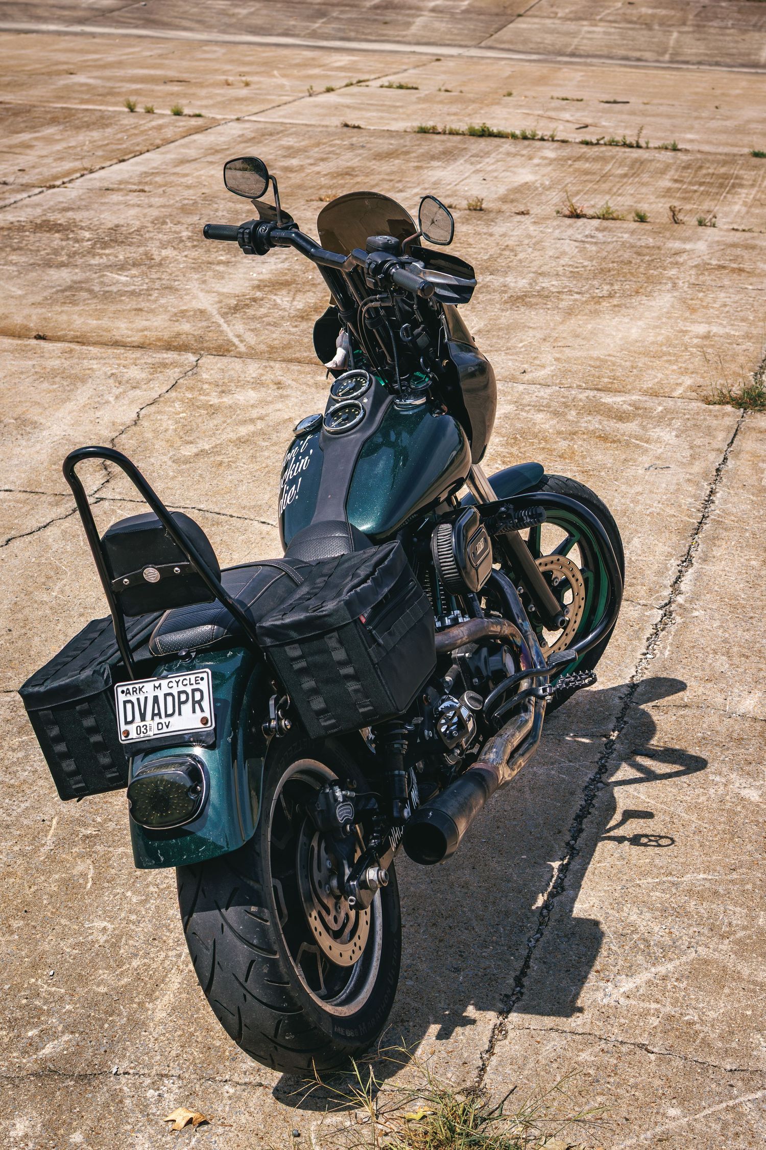 Dark green custom Harley-Davidson motorcycle with saddlebags parked on concrete, rear view showing license plate DVADPR.