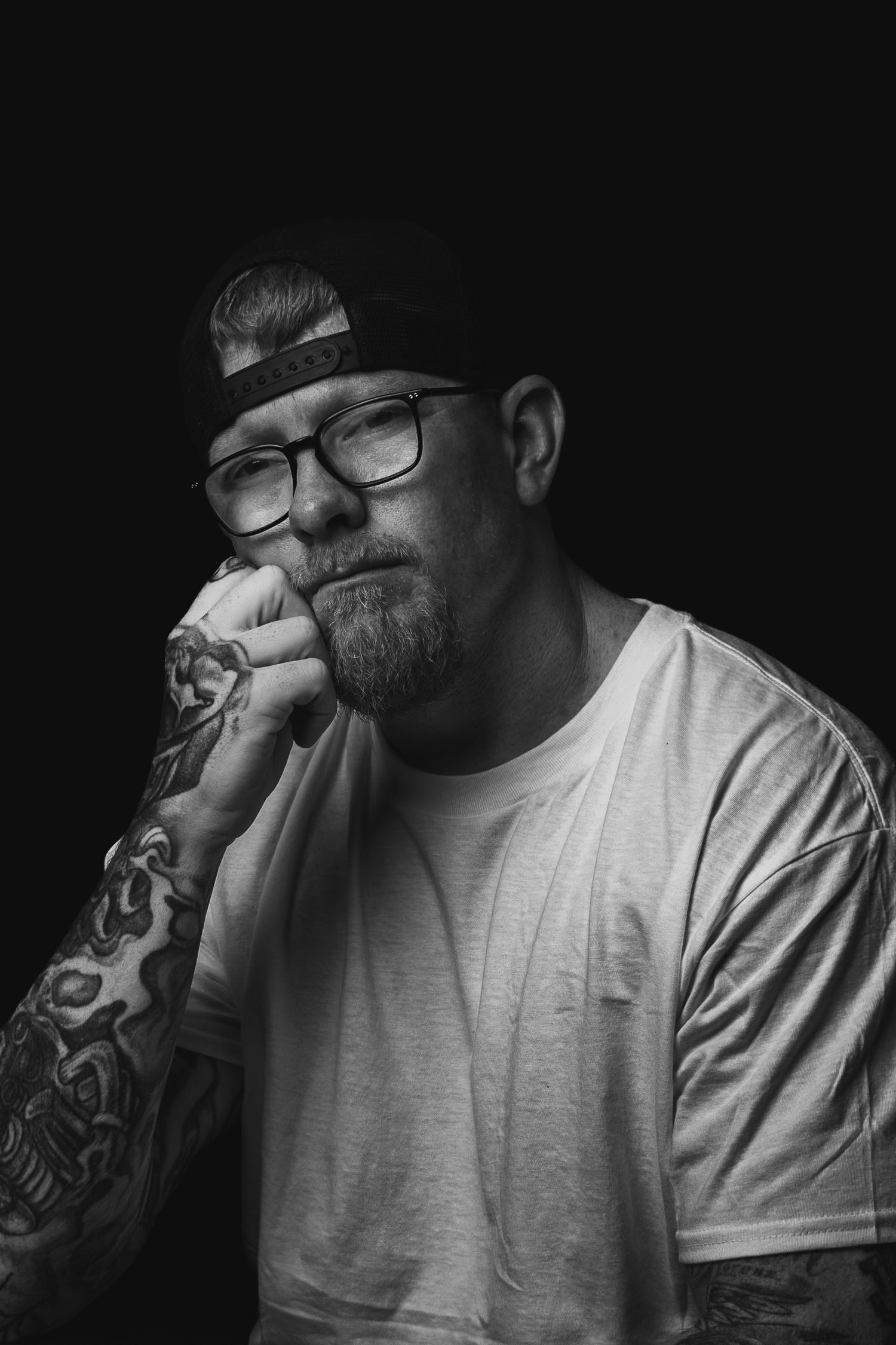 Black and white portrait of a bearded tattooed man with glasses resting his fist against his cheek.