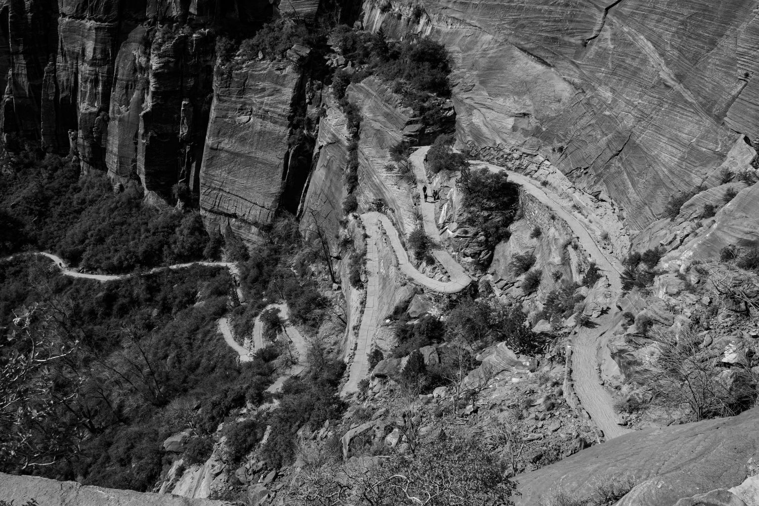 Black and white aerial view of a winding switchback trail carved into rocky canyon cliffs.