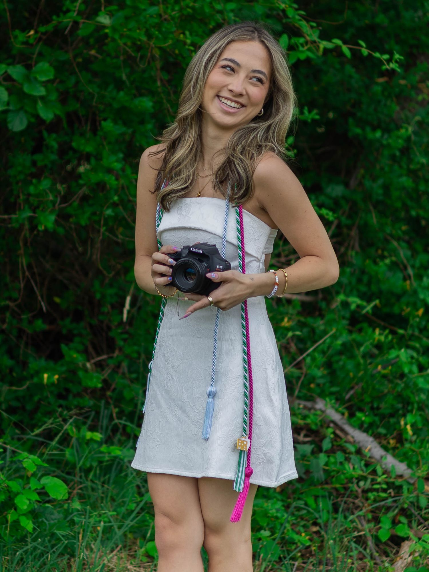 Smiling young woman in white dress holding a camera with colorful strap outdoors.