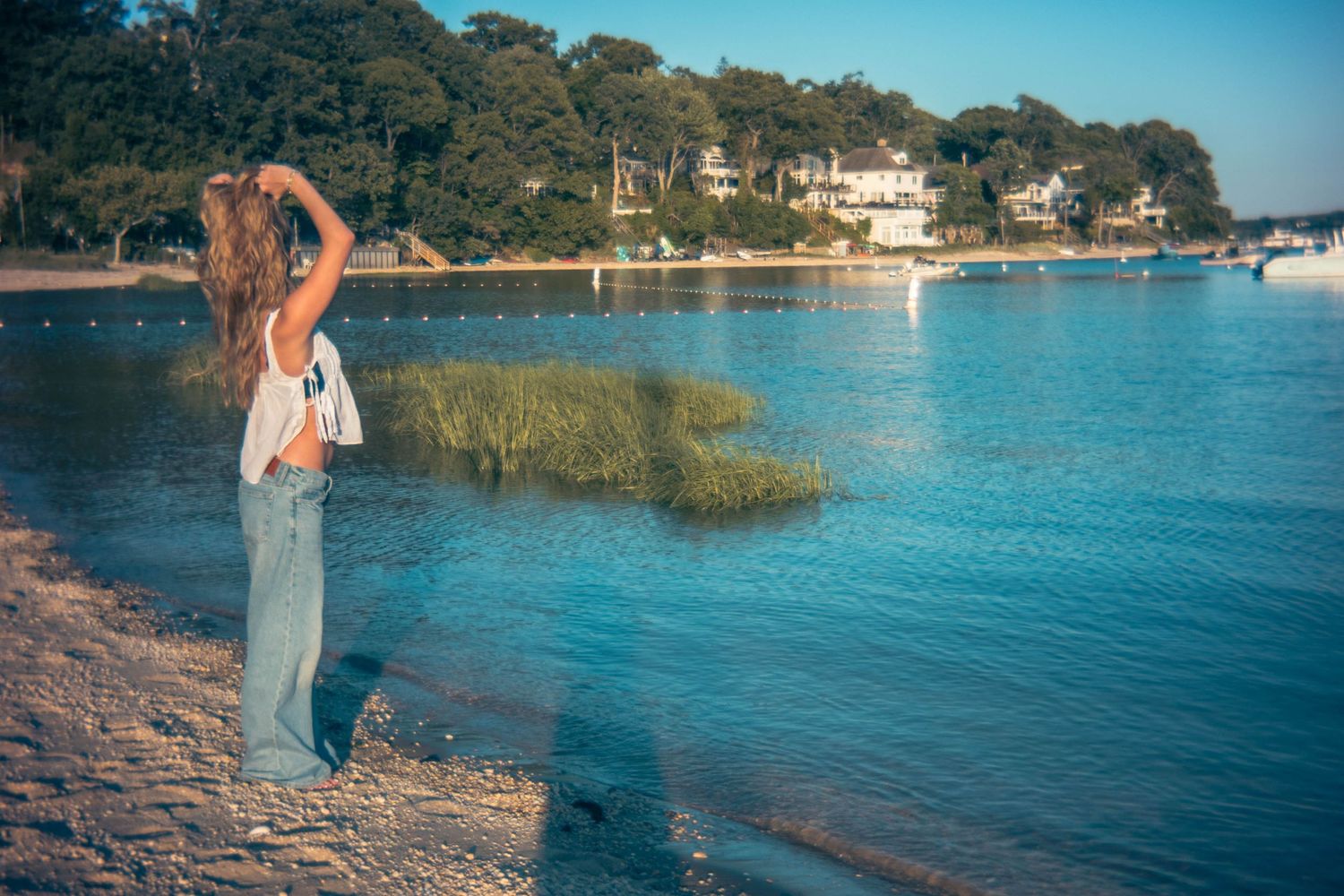 Woman in jeans standing on sandy shore gazing at calm blue water with lush trees in background.