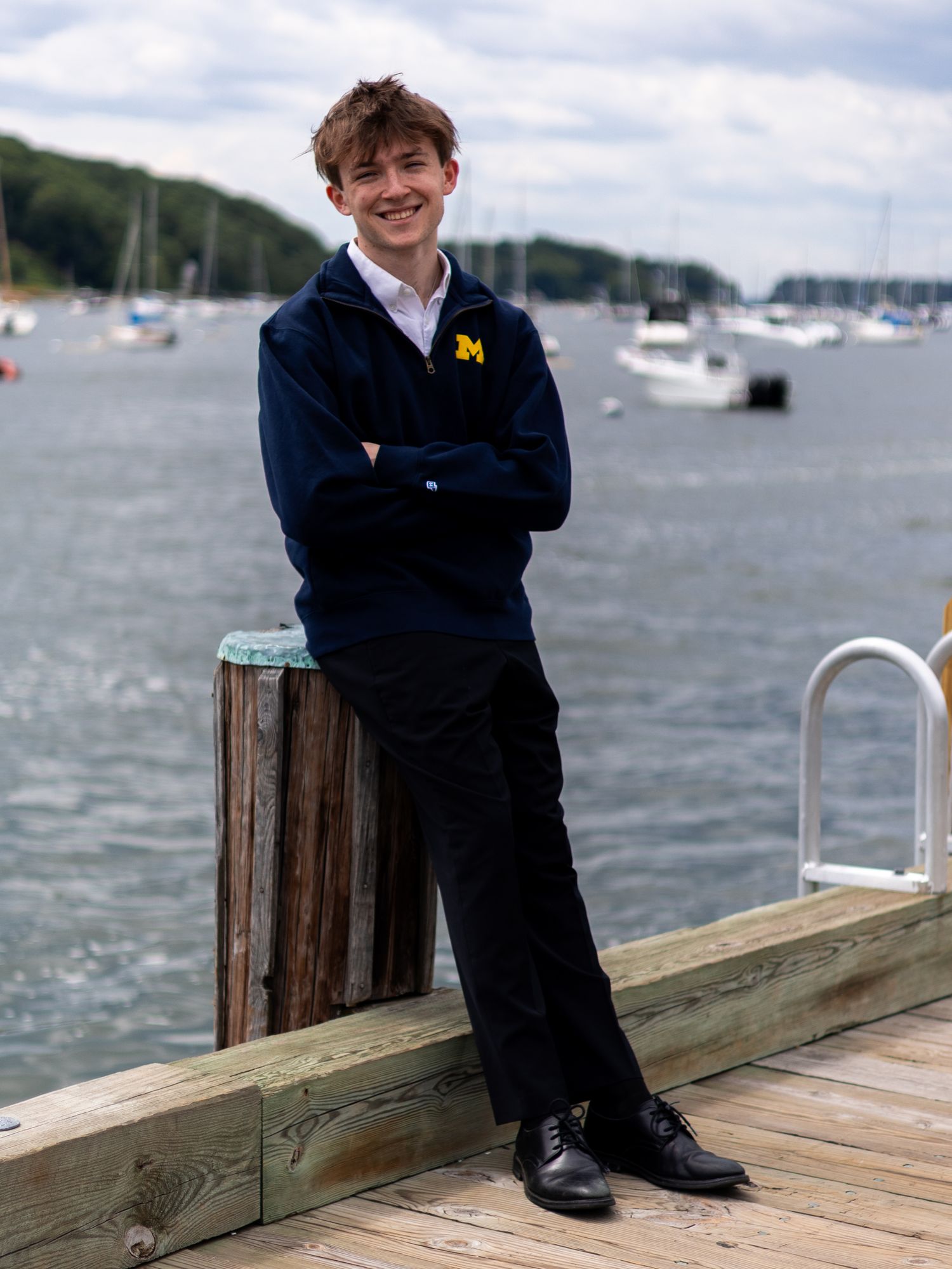 Smiling teen in navy Michigan pullover posing on wooden dock with harbor behind him.