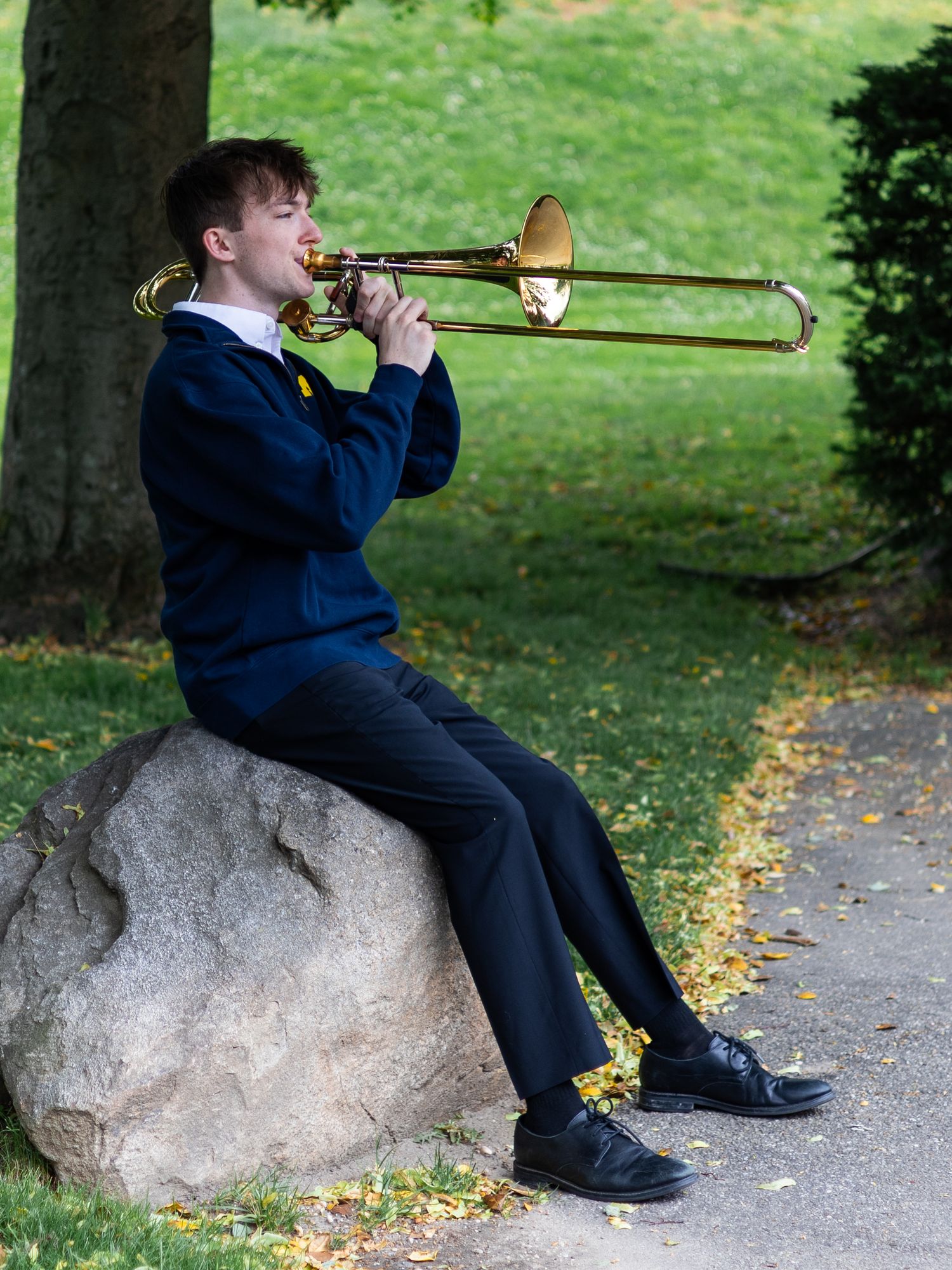 Young musician playing trombone while seated on large rock in green park setting.
