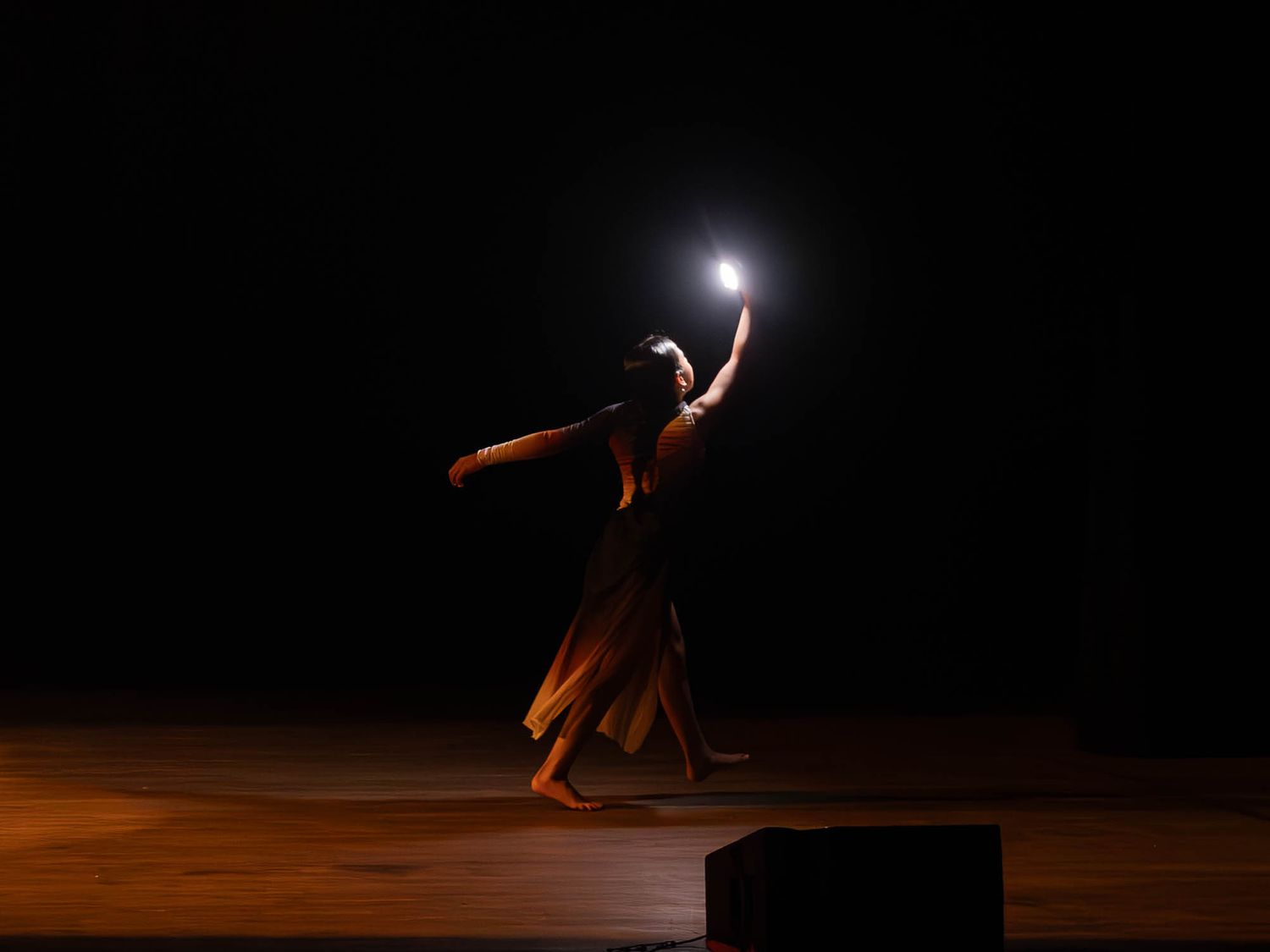 A solo dancer in a flowing brown dress reaches toward a bright light on a dark theatrical stage.