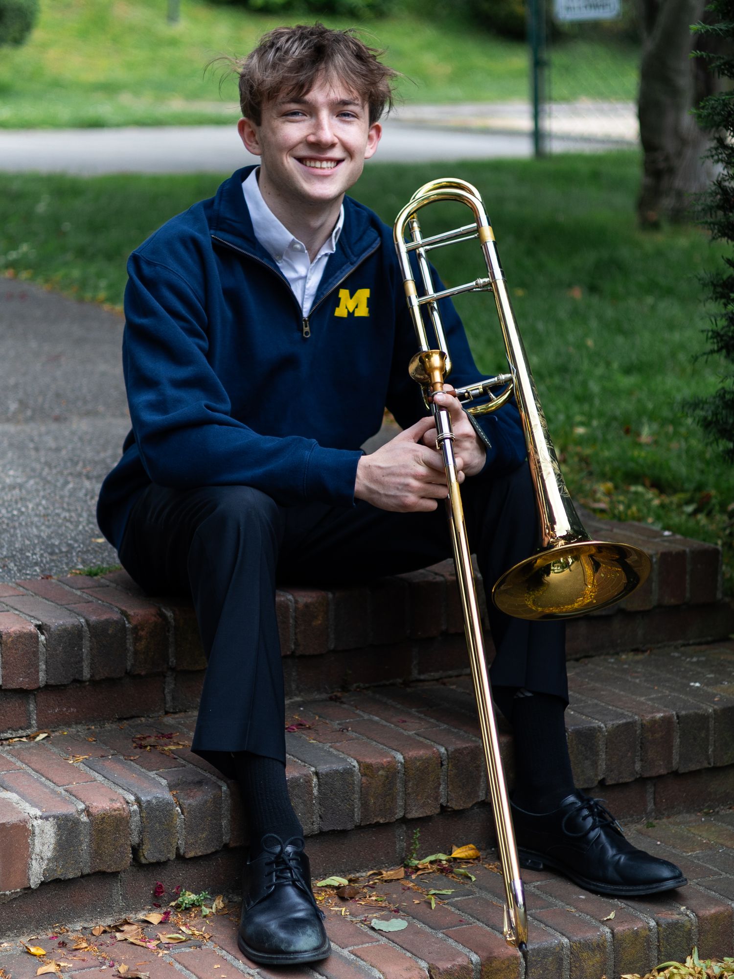 Smiling young man in a navy Michigan jacket sits on brick steps holding a trombone outdoors, with fallen leaves and green grass behind him.