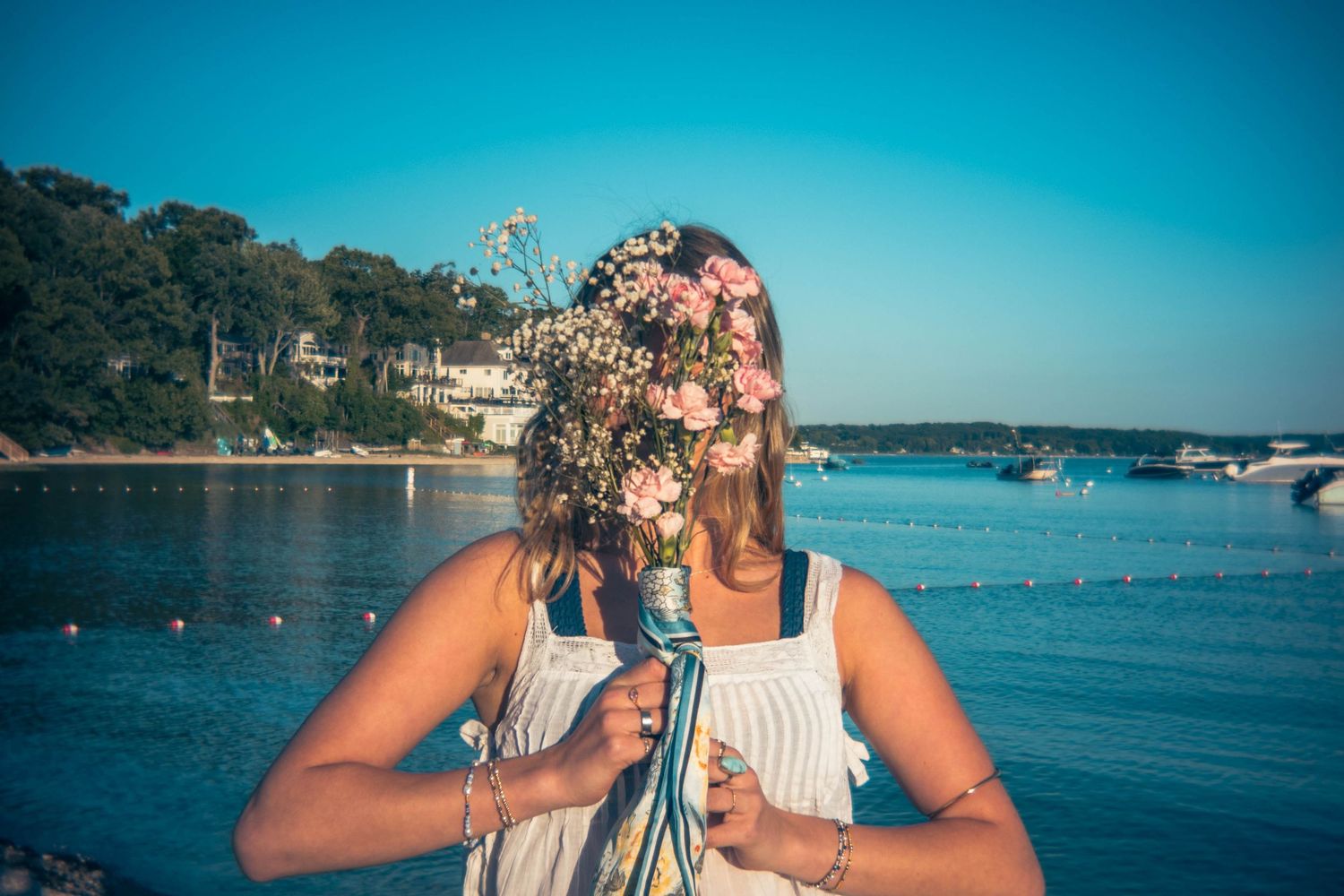 Woman in a white top holds a bouquet of pink wildflowers over her face beside a calm turquoise bay with boats and a wooded shoreline.