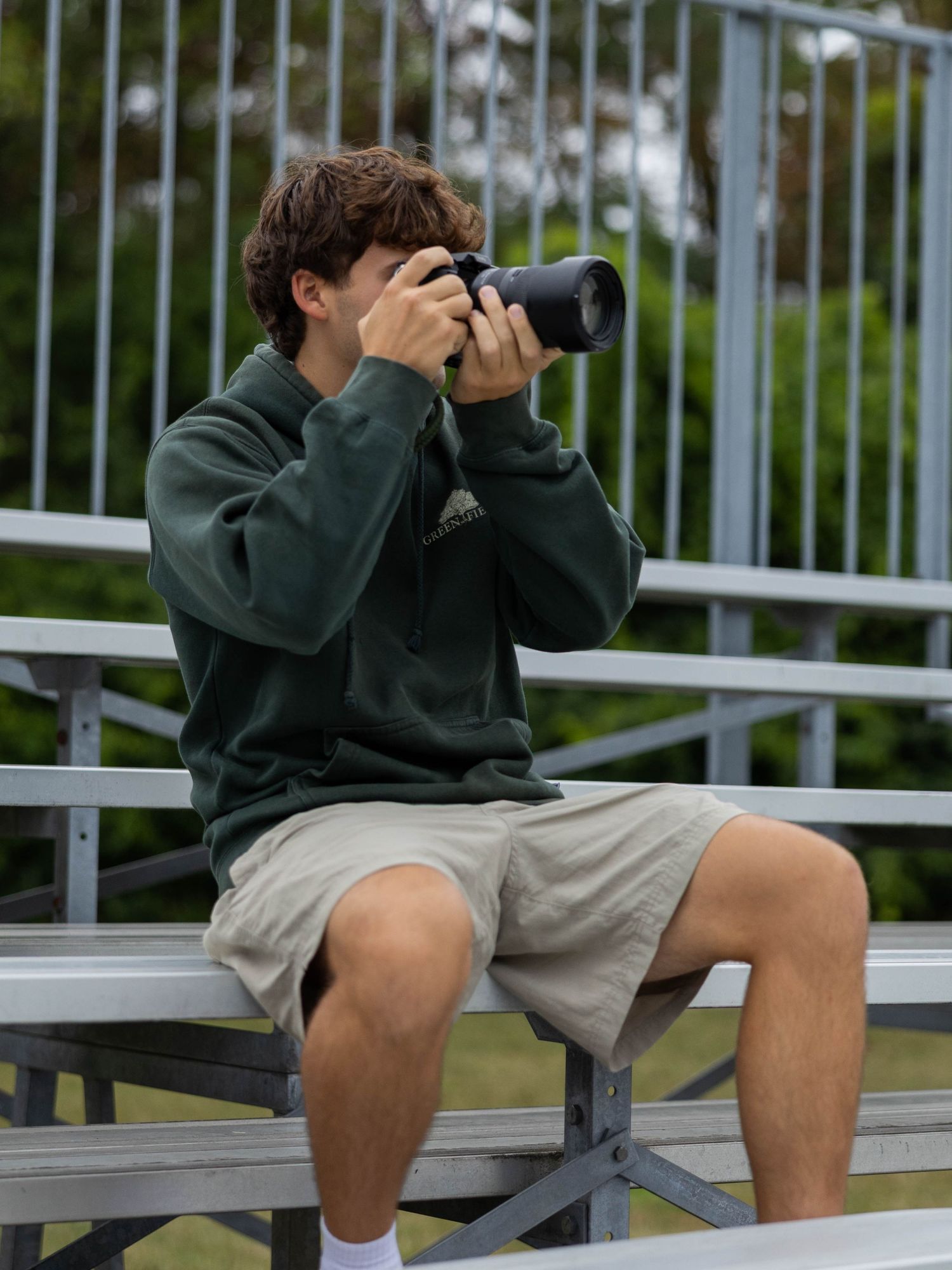 A young man in a green hoodie and khaki shorts sits on bleachers, taking photos with a DSLR camera outdoors.