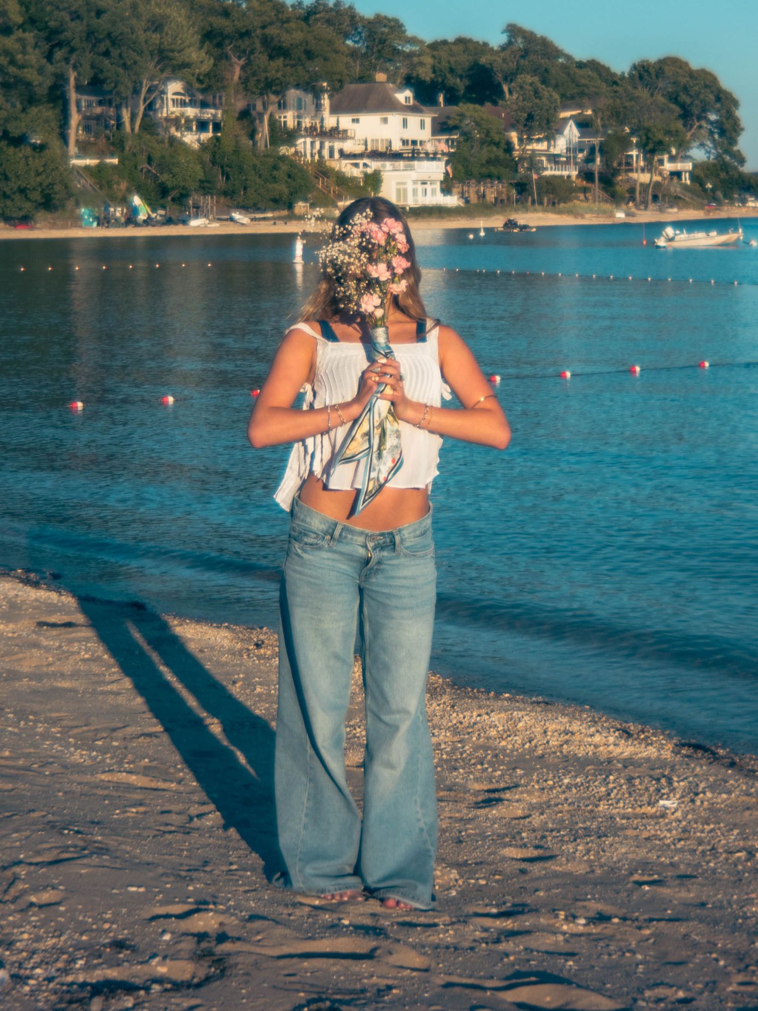 Woman in white crop top and wide-leg jeans holding flowers at sandy beach with blue water.