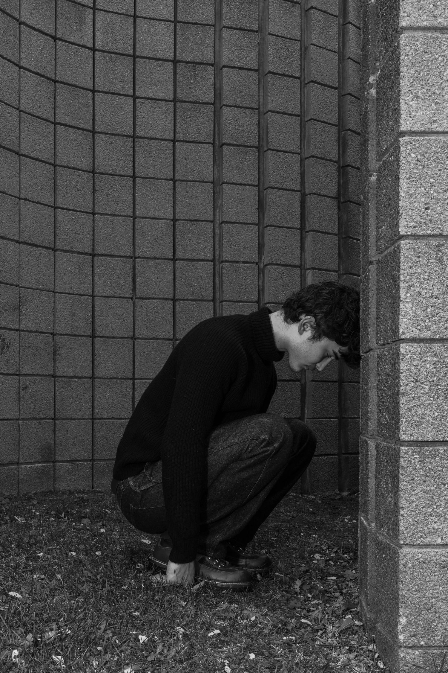 Young man in a dark sweater crouches with his forehead pressed against a concrete block wall in a black and white photo.