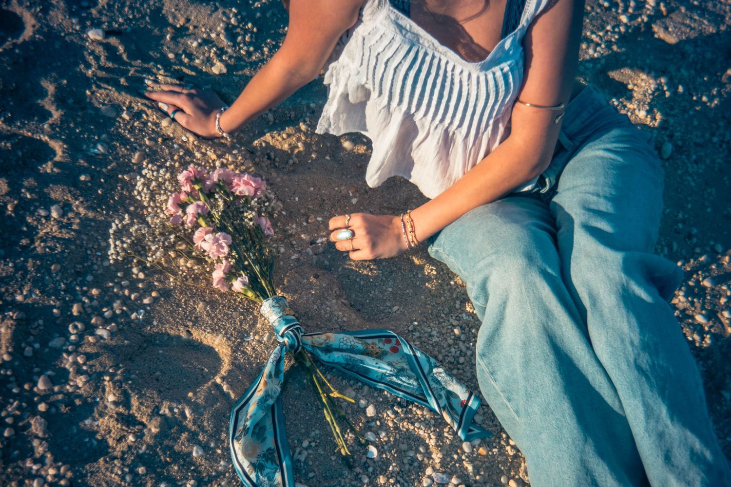Woman in a white ruffled top and light blue jeans sits on a pebbly beach at golden hour, resting her hands beside a pink flower bouquet.