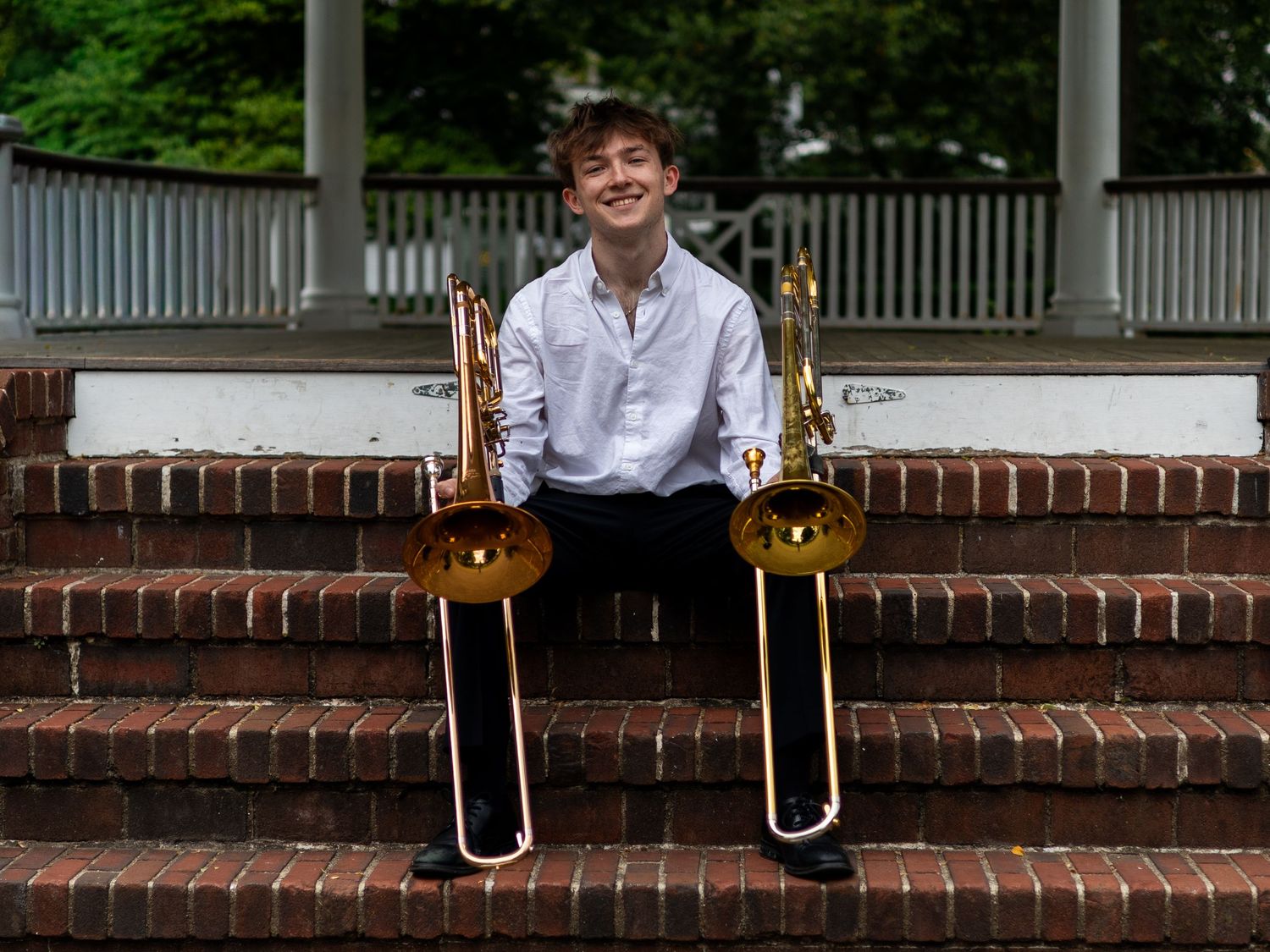 Young musician smiling on brick steps holding two trombones outdoors near a gazebo.