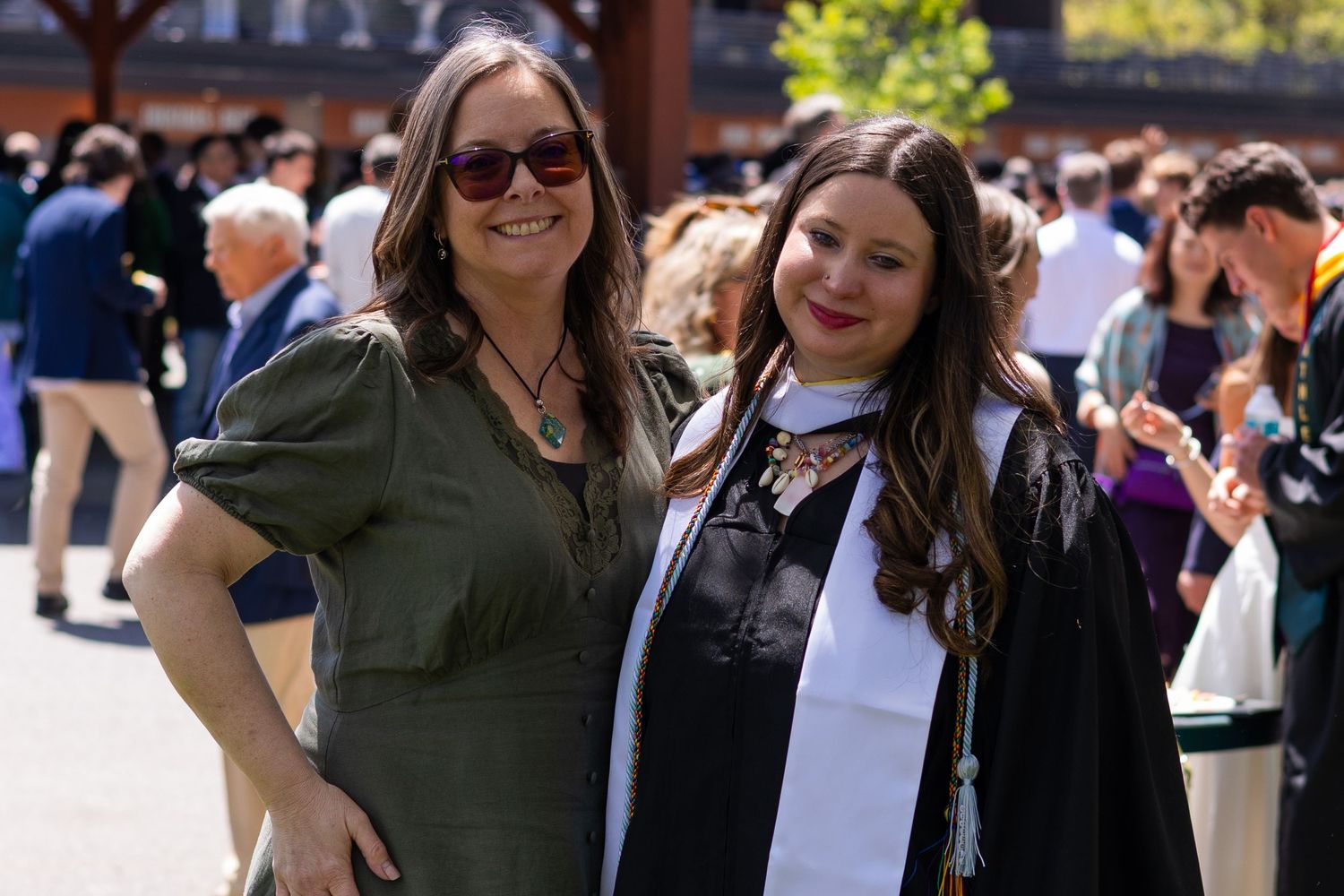 Two women smile at a graduation ceremony, one wearing a cap and gown with a white stole outdoors.