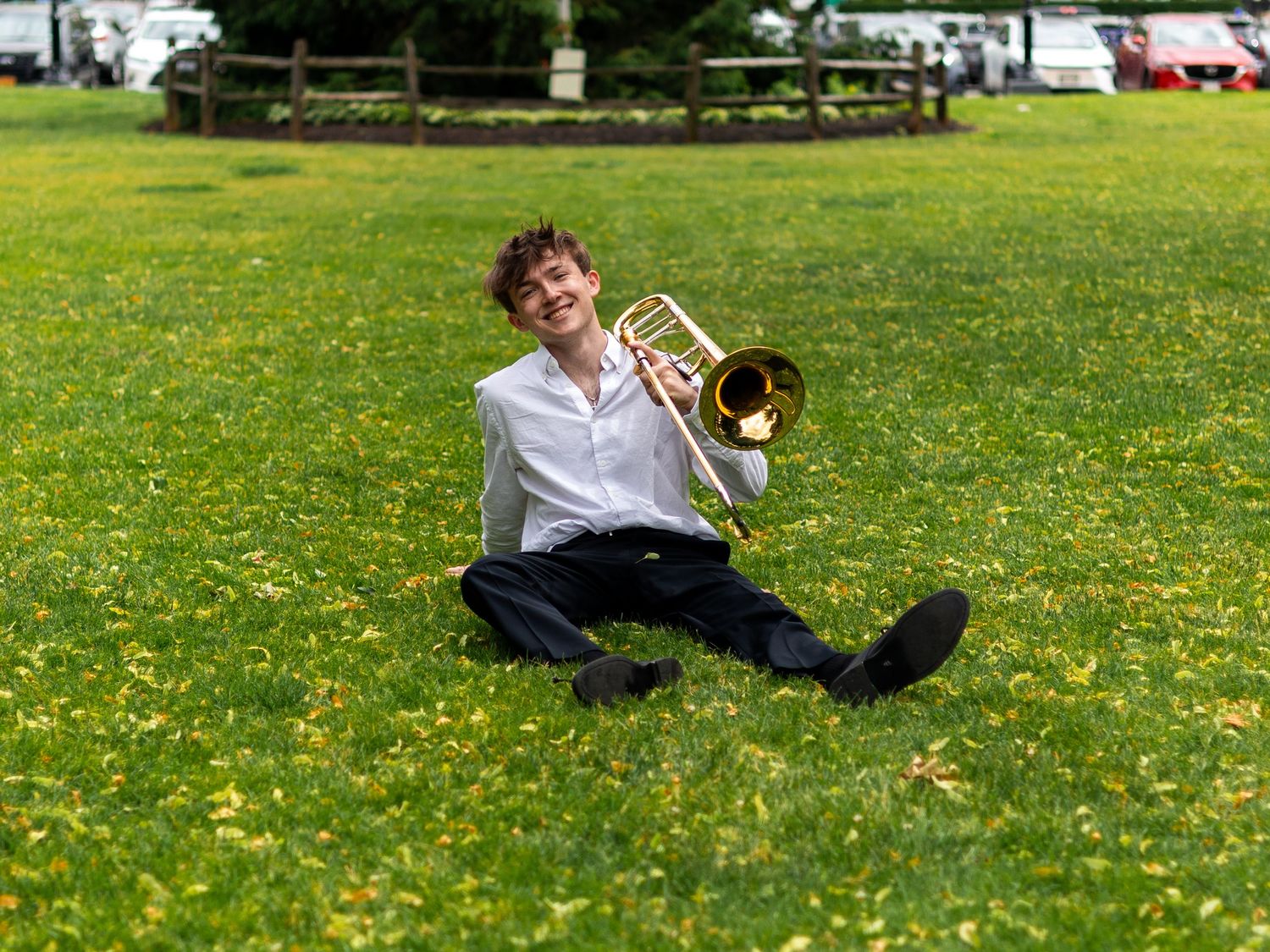 Young musician sitting on grass laughing while holding trombone in sunny green field.