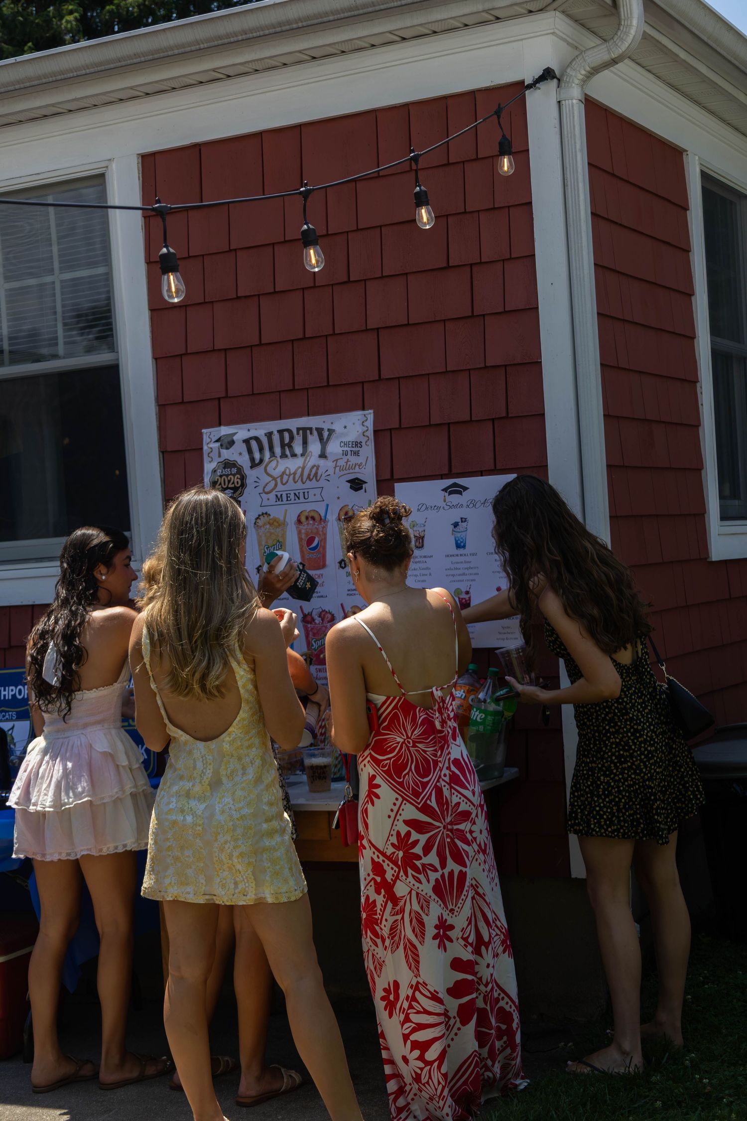 Group of young women browse a Dirty Soda menu display outside a red house at a summer party.