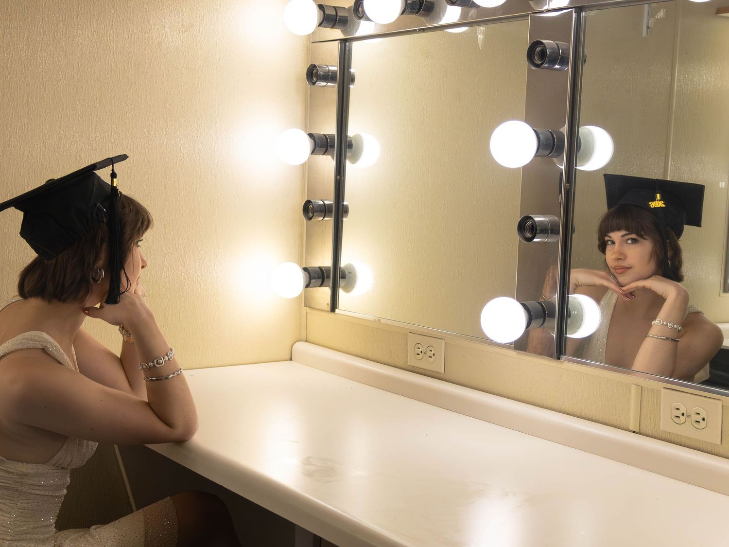 A graduate in cap poses at a Hollywood-style vanity mirror with bright bulb lights in a dressing room.