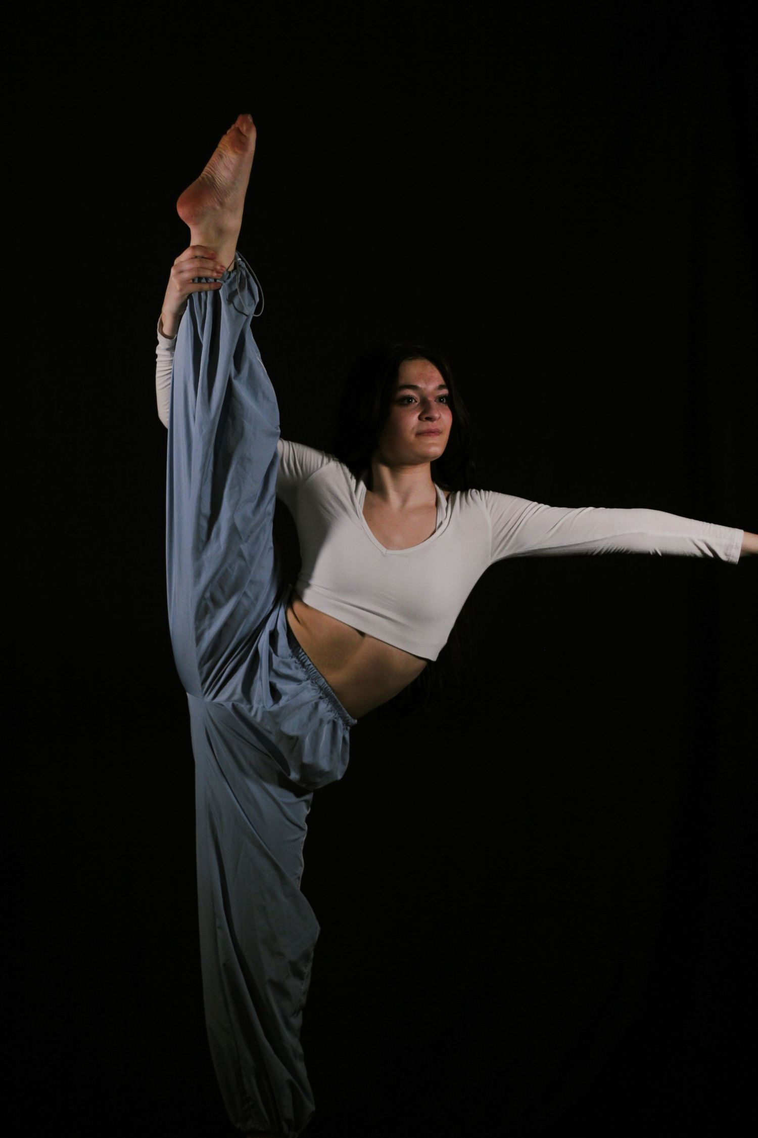 Dancer in a white crop top and flowing blue pants performs a standing vertical split against a dark studio background.