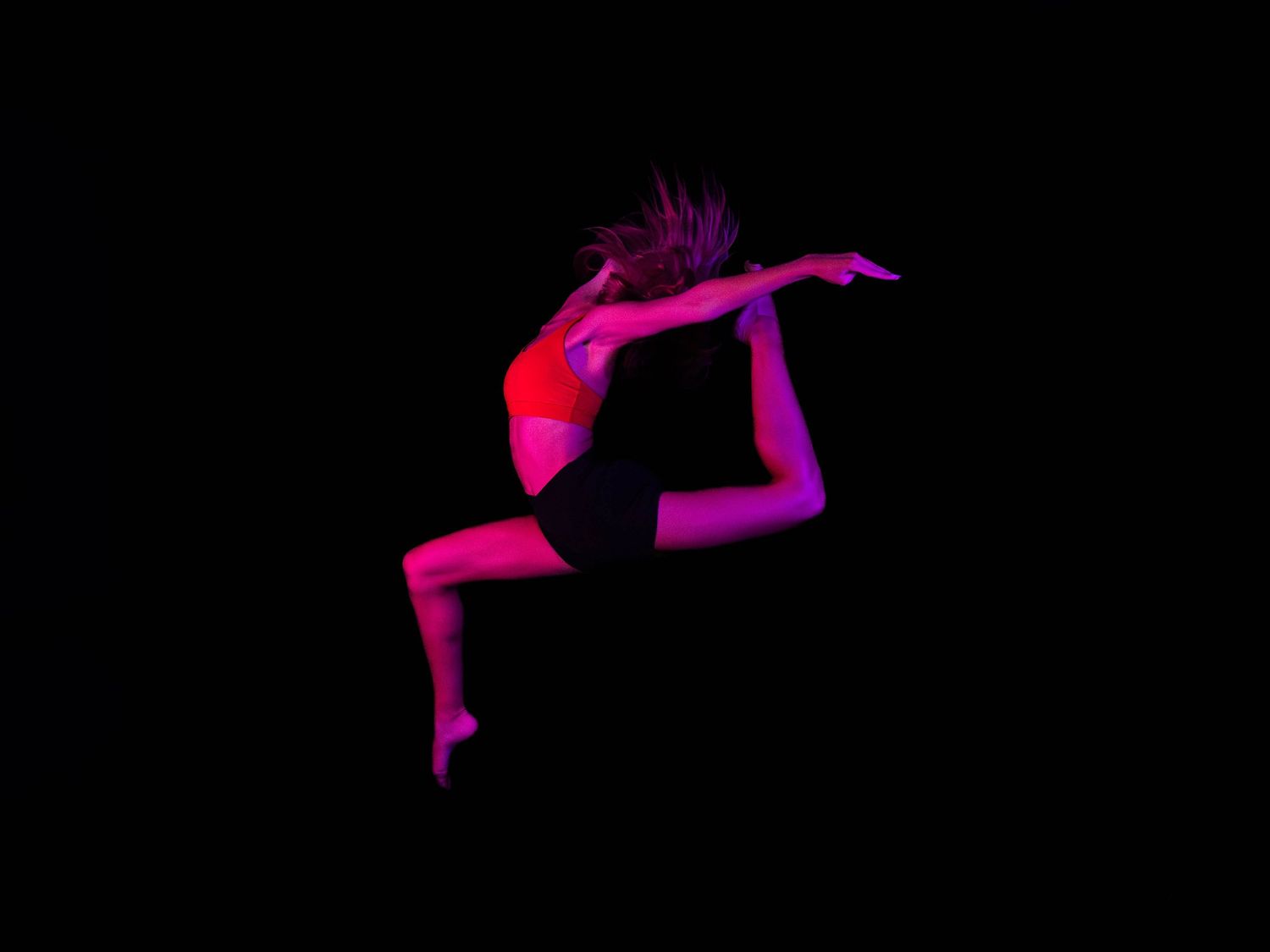 Dancer in a red top and black shorts leaps mid-air in a graceful split jump, lit by vivid pink and magenta stage lighting against a black ba