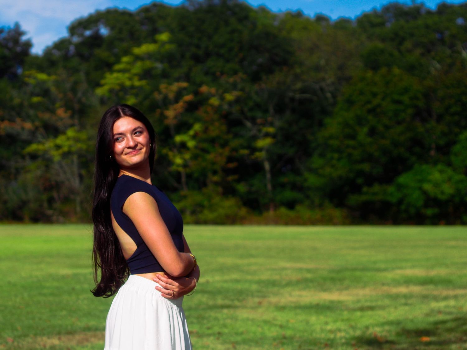 Brunette woman in navy top and white skirt smiling over shoulder in lush green field on sunny day.